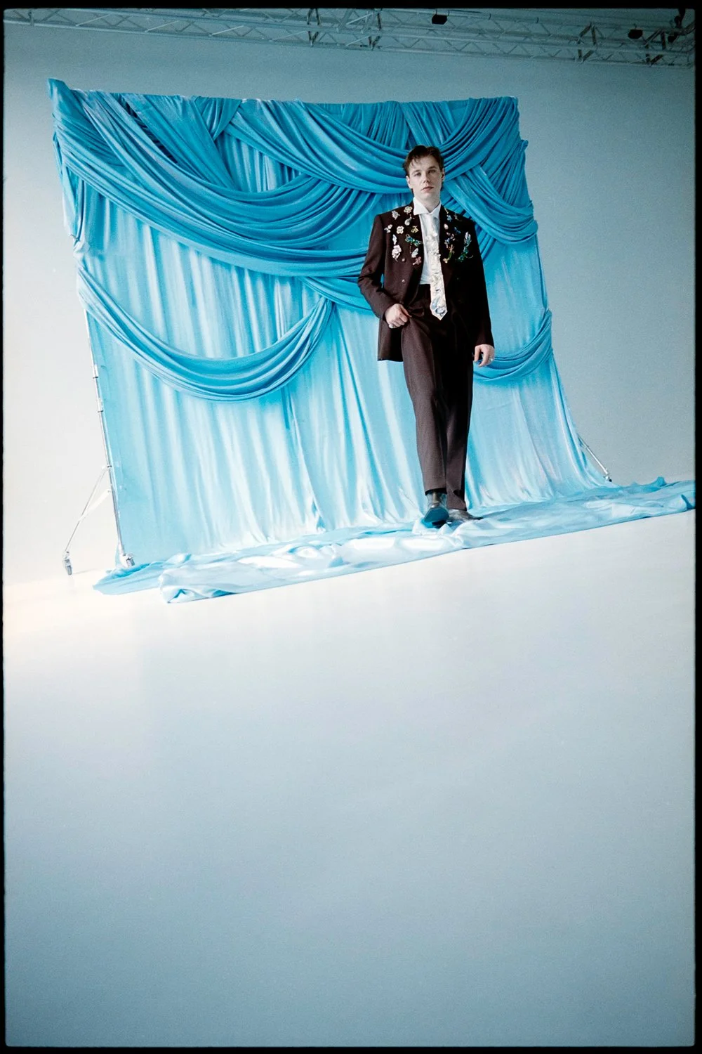 A person walking on stage with a blue, draped backdrop, wearing a dark suit with embroidered designs and a light-colored shirt.
