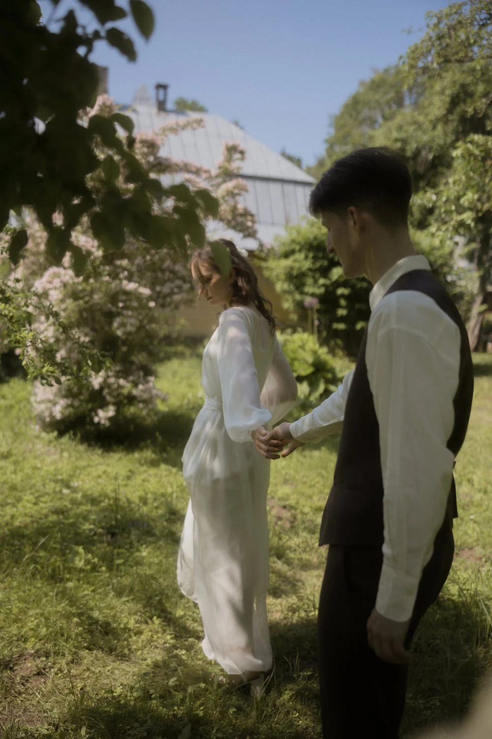 A man and woman holding hands outdoors in a lush, green garden with a house and trees in the background.