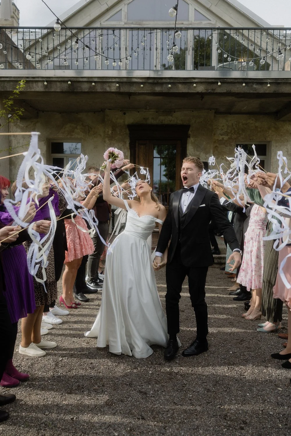 Wedding celebration with a bride and groom holding hands, walking through a ribbon-cutter line of guests outside a rustic building, with a woman throwing flower petals and string lights overhead.