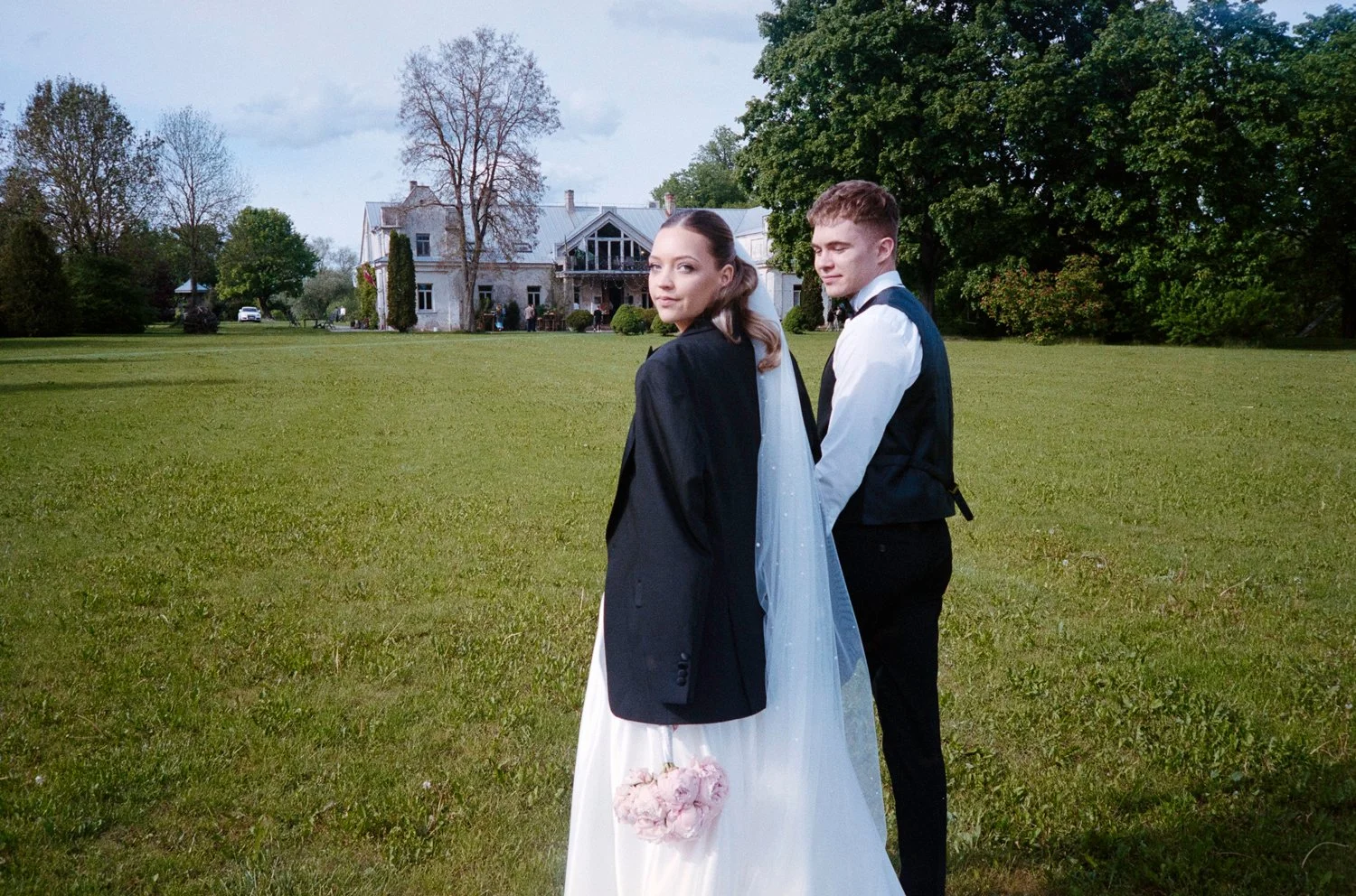 A bride and groom standing on a large green lawn with a large house and trees in the background.