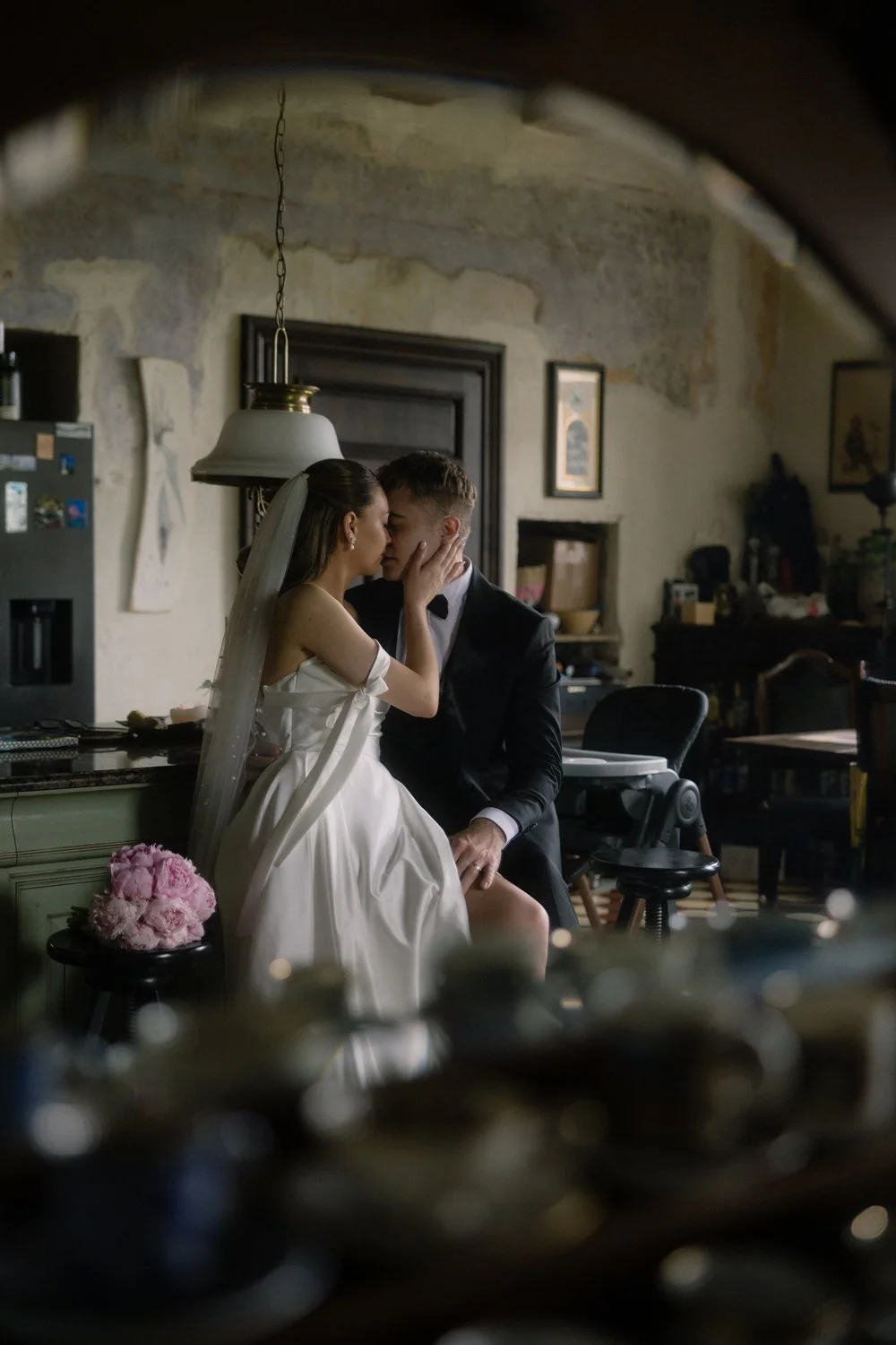 A bride and groom share an intimate moment in a rustic room, sitting close and touching foreheads, with the bride wearing a white wedding dress and veil, and the groom in a black tuxedo.