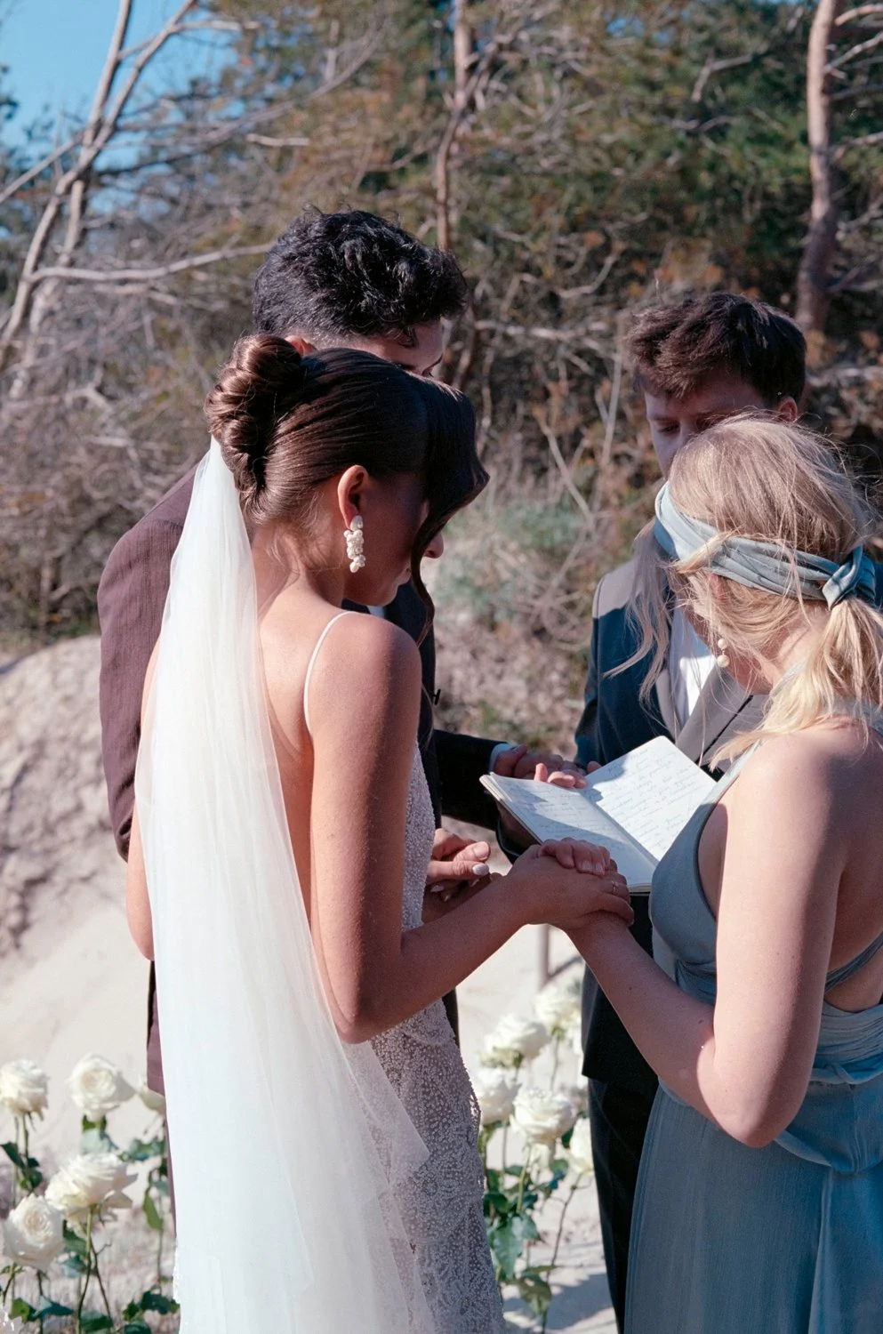 A bride and groom standing outdoors during a wedding ceremony, holding hands and exchanging vows, with a woman reading from a book and two men observing, surrounded by white flowers and natural scenery.