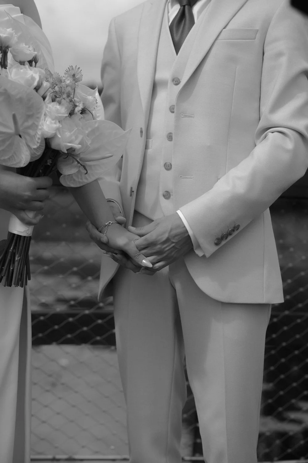 A groom in a light-colored suit holding hands with a bride in a dress holding a bouquet of flowers.