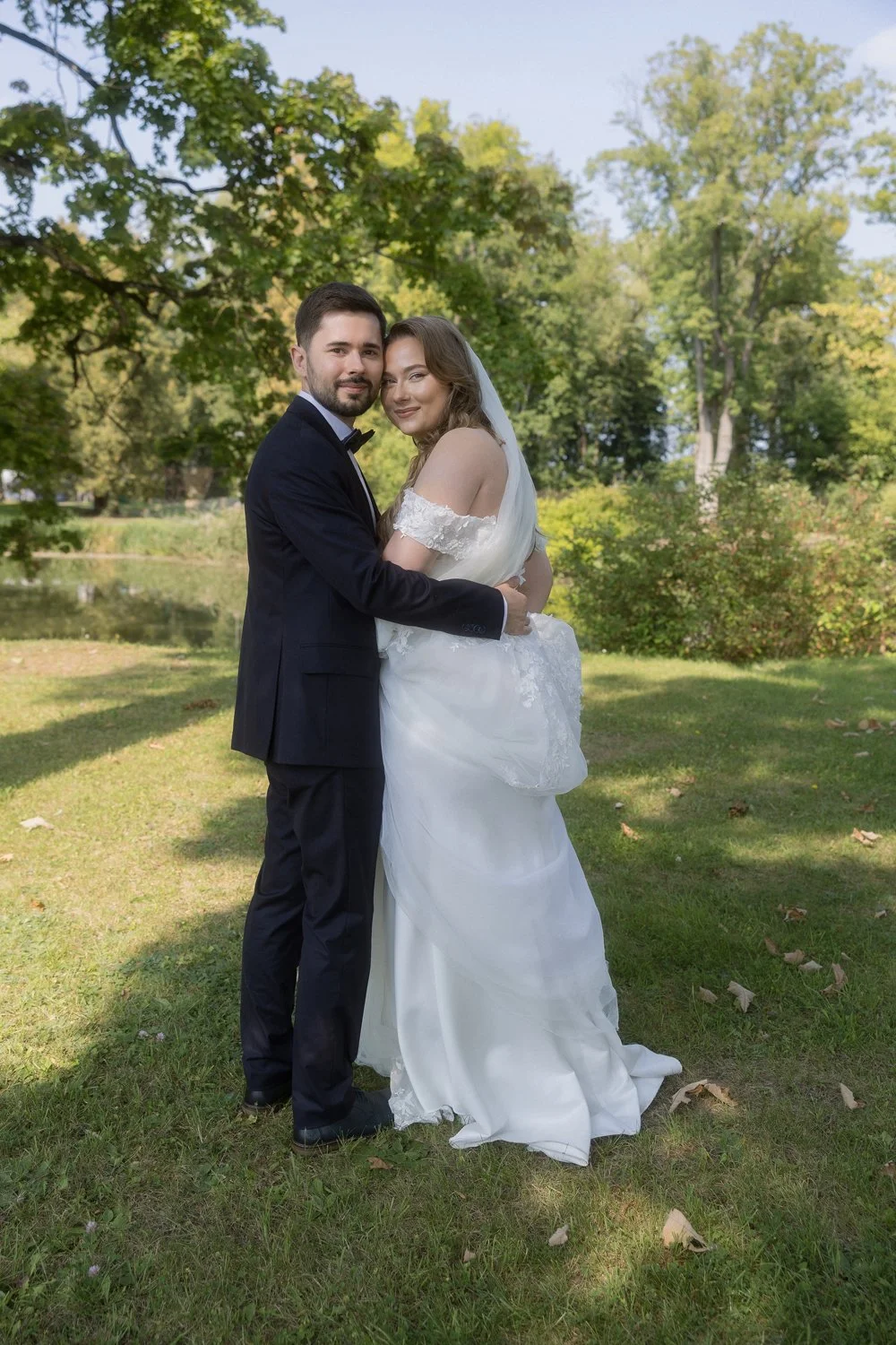 A newlywed couple standing outdoors on a grassy area near a pond, surrounded by trees, on a sunny day.