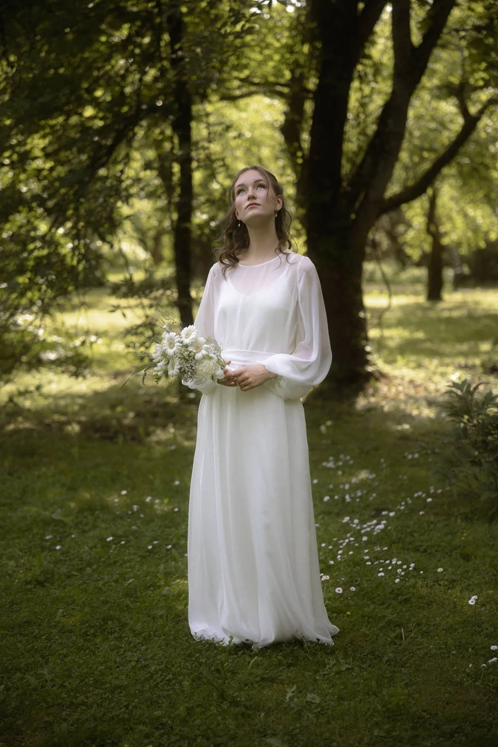 A woman in a white dress holding a bouquet of white flowers, standing in a green outdoor setting with trees and grass.