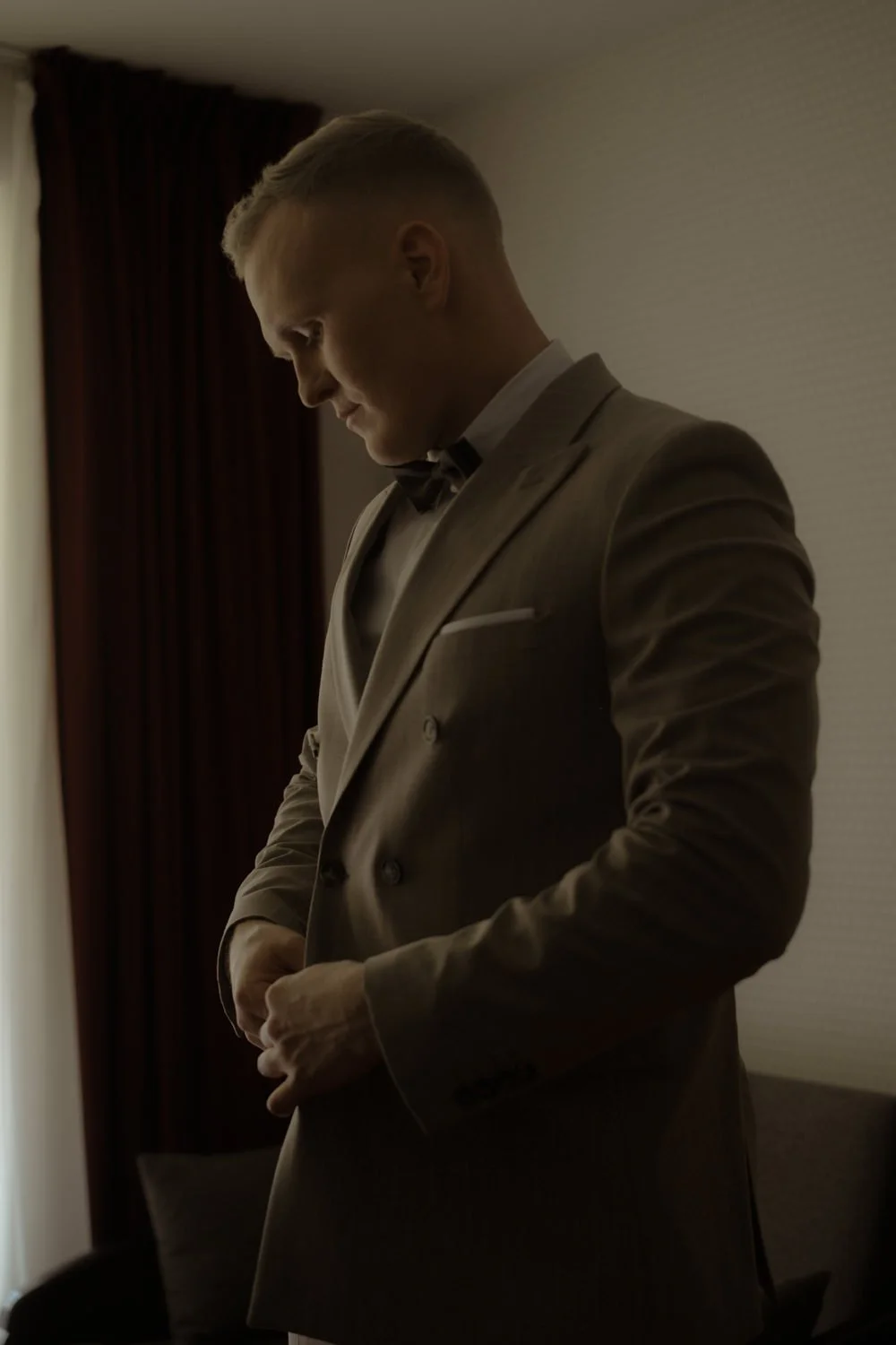 A man in a light-colored tuxedo adjusting his sleeves in a dimly lit room.