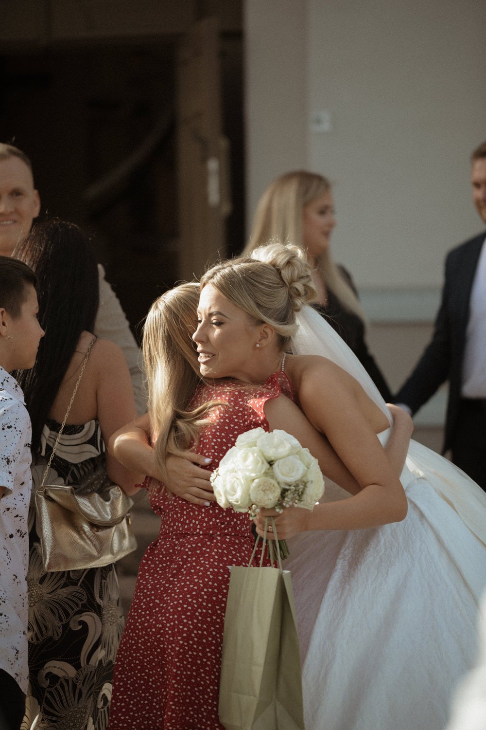 A bride in a white wedding dress hugging a woman in a red dress, who is holding a white bouquet and a gift bag, while family members surround them at a wedding celebration.