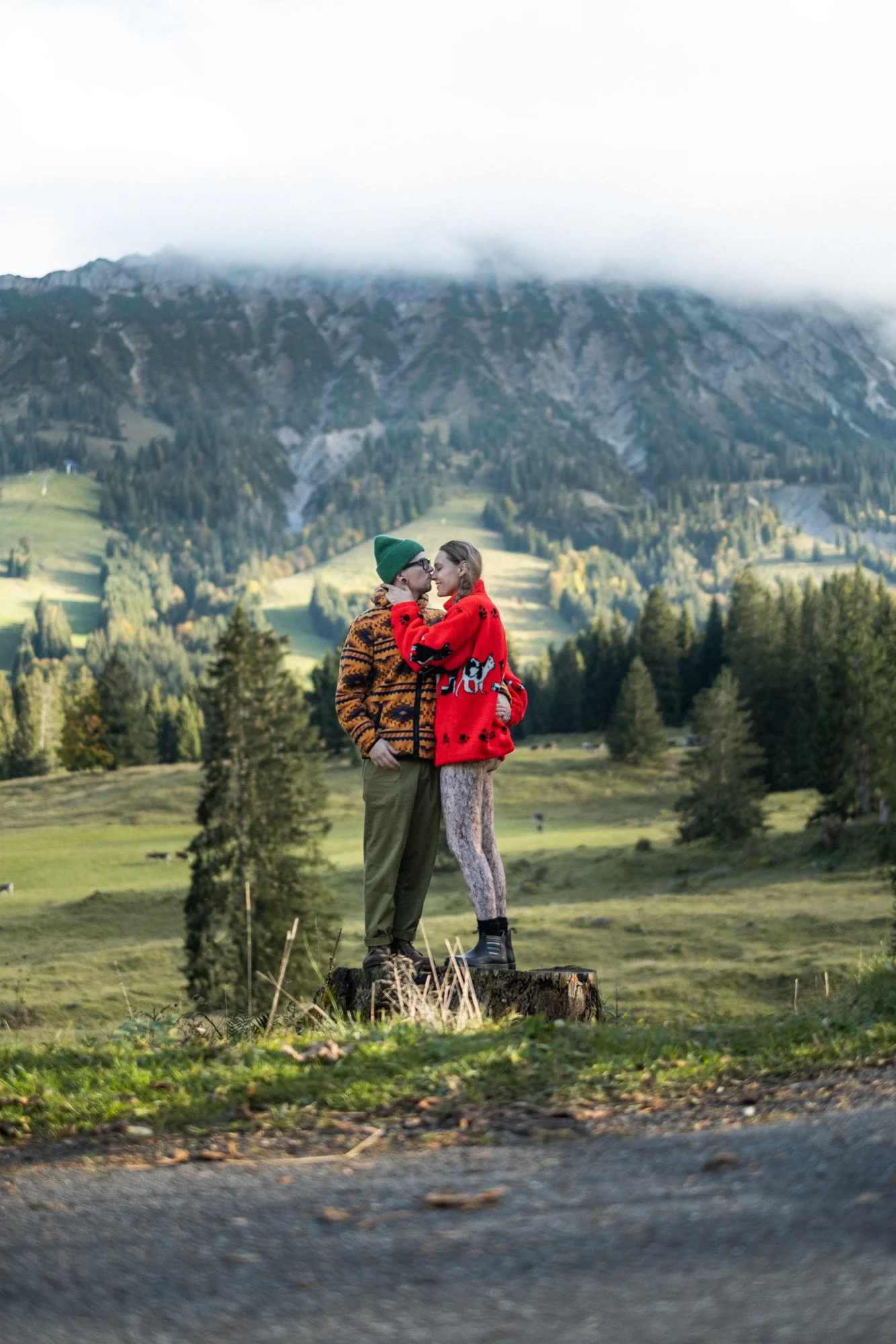 A couple standing on a tree stump in a scenic mountain landscape, wearing colorful winter clothing, with mountains and trees in the background.