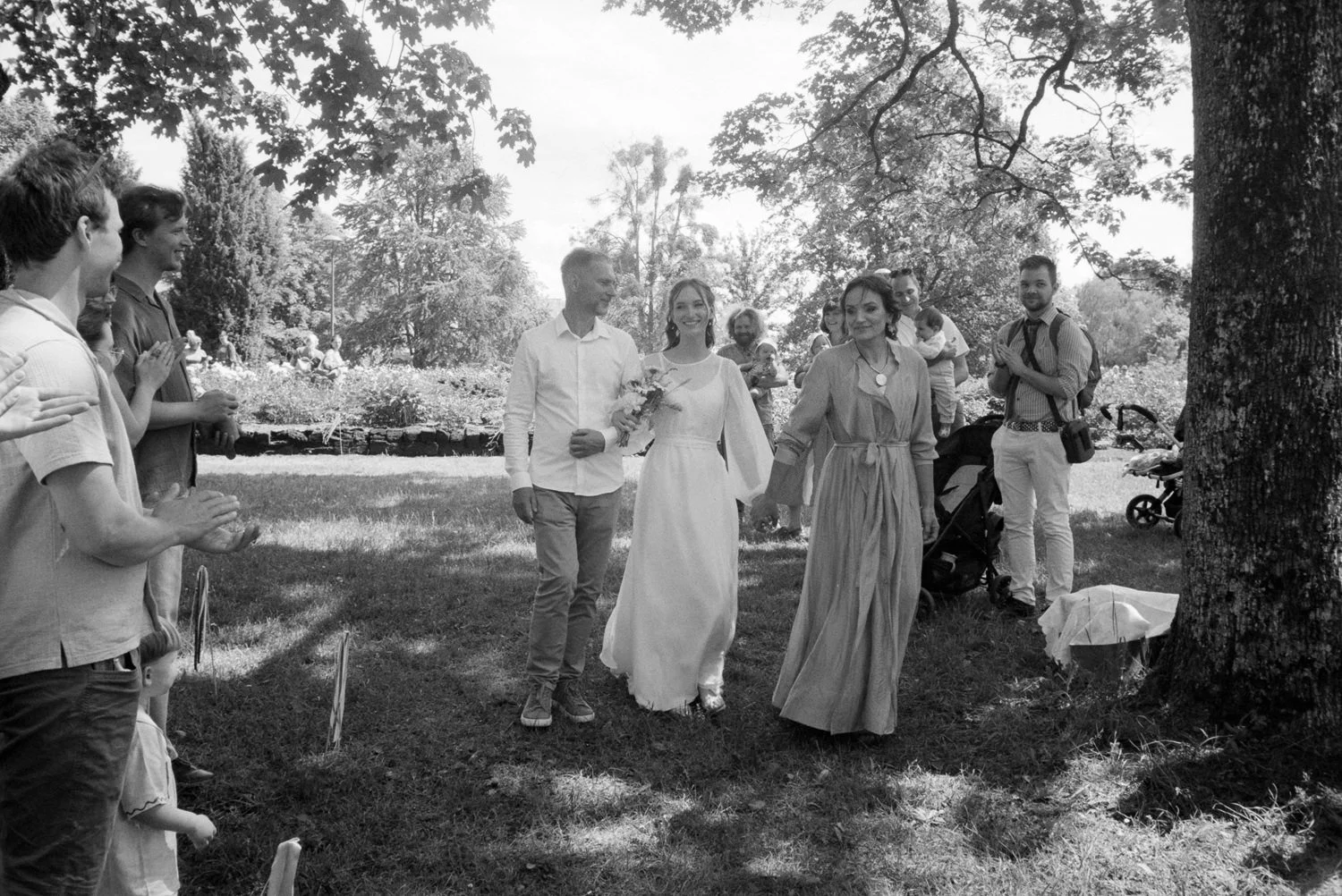 A bride and groom walking hand in hand through a park gathered with friends and family, who are clapping and smiling.