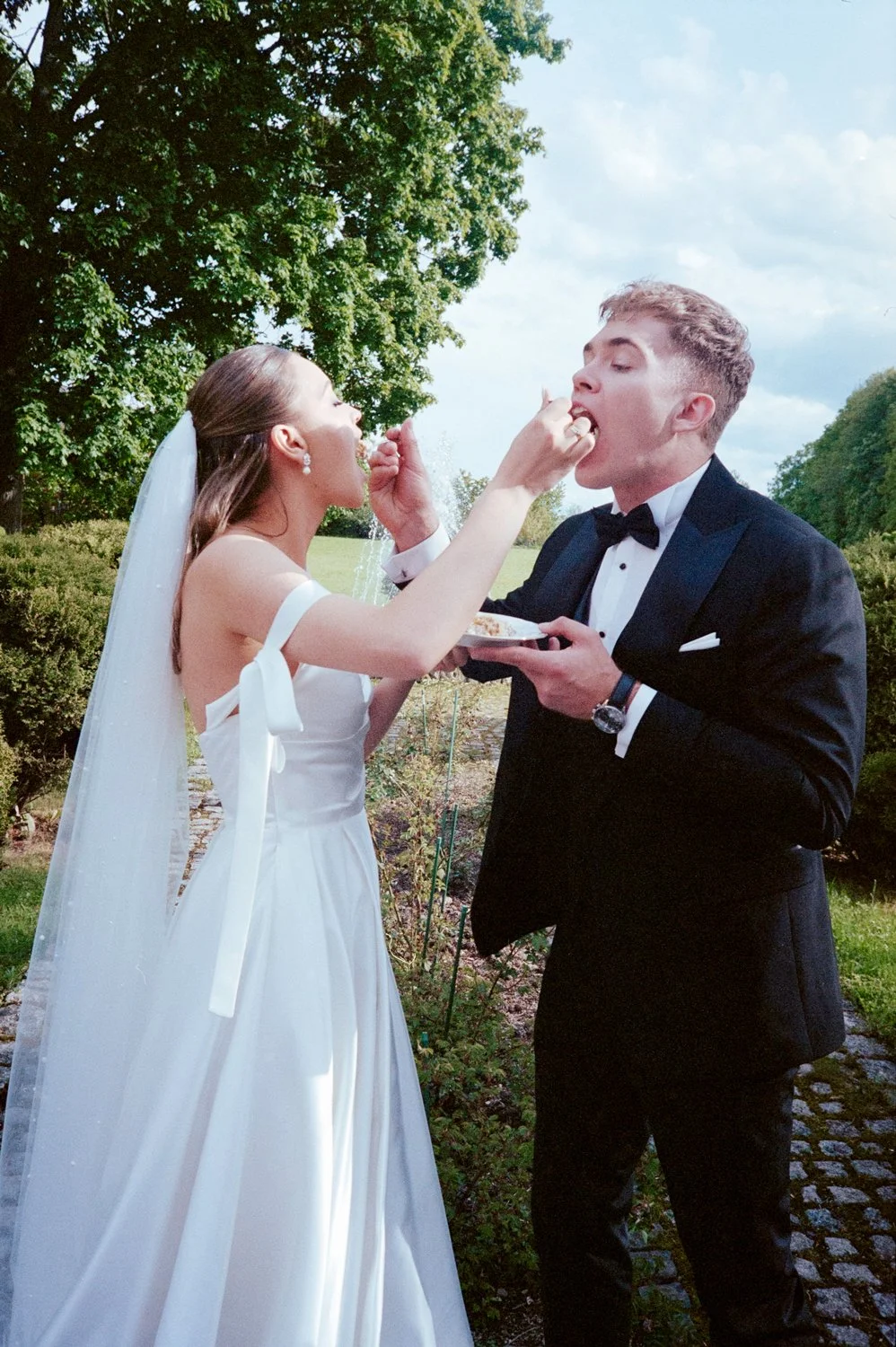 Bride in a white wedding dress with veil and earrings sharing cake with groom in a black tuxedo outdoors with green trees and cloudy sky in the background.