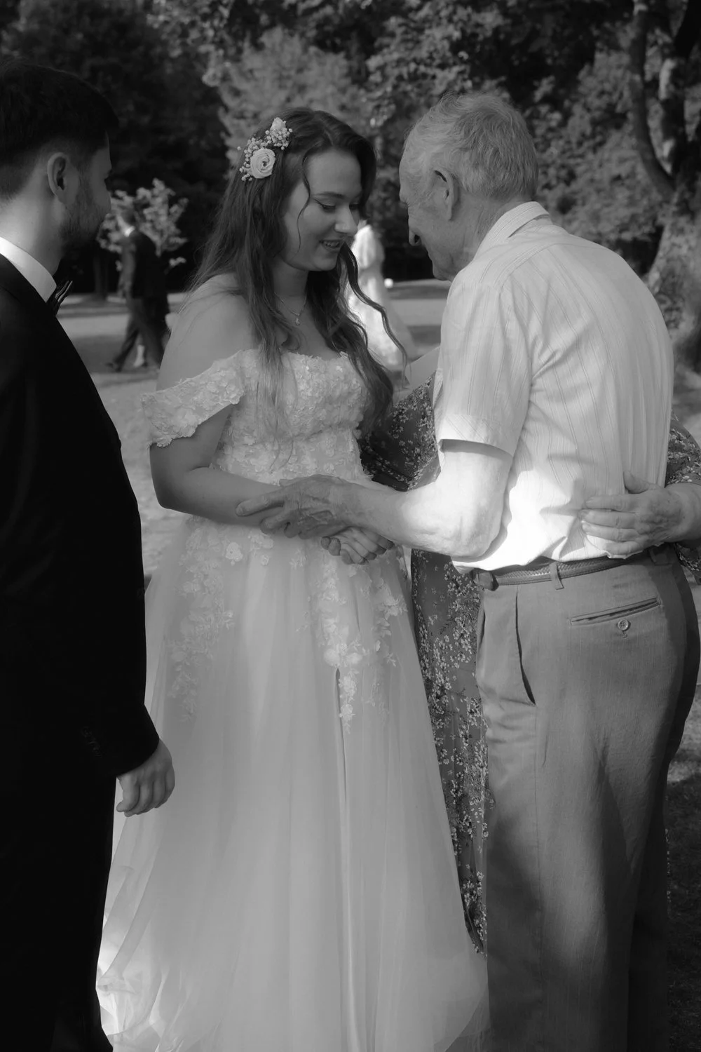 An elderly man shaking hands with a young woman in a wedding dress during a wedding ceremony outdoors, with a groom standing nearby.