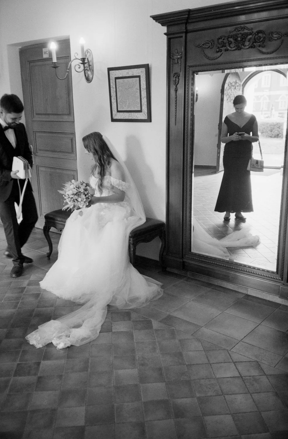 A bride in a wedding dress sitting on a bench holding a bouquet, with a man standing nearby in formal attire. A woman in a long black dress is reflected in a large ornate mirror, looking at her phone, with the entrance door open behind her showing th