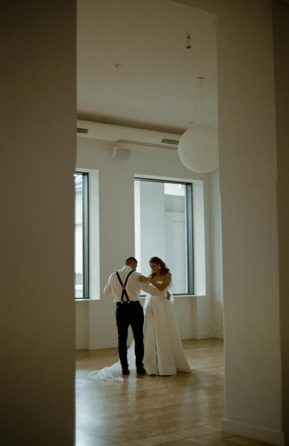 A bride and groom dancing in a minimally decorated room with large windows and natural light.