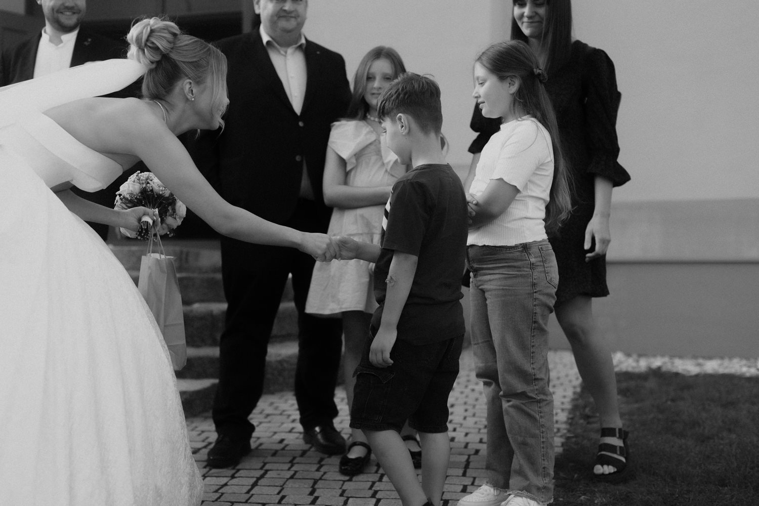 A woman in a wedding dress shaking hands with a young boy, with group of people watching in the background.