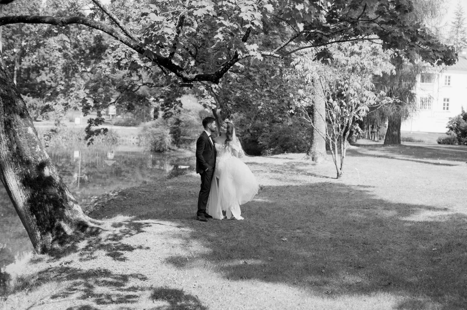 Black and white photo of a bride and groom standing under a tree near a pond, smiling and looking at each other in a park setting.