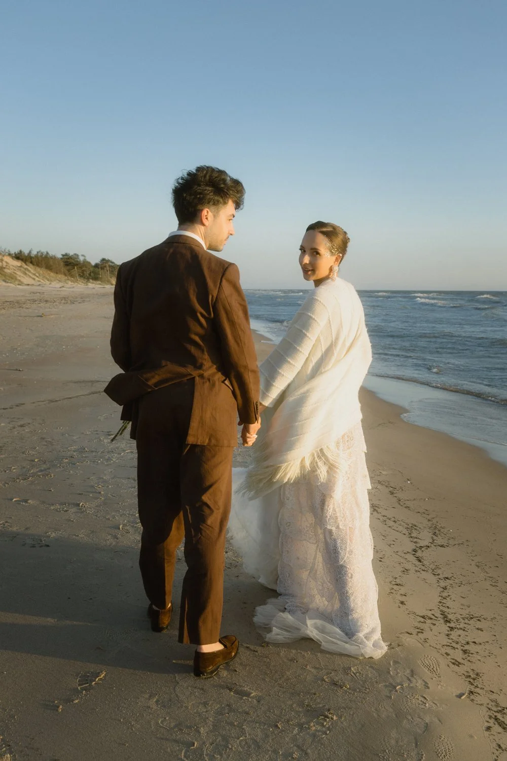 A couple holding hands and walking on the beach, dressed in wedding attire, during sunset or late afternoon.