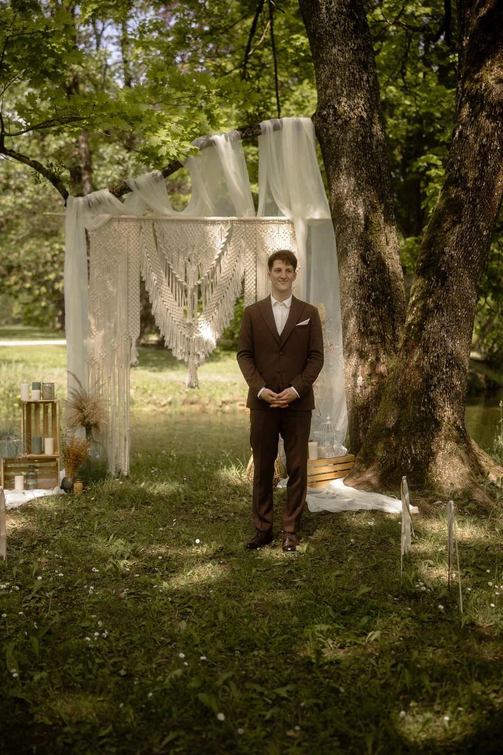 A young man in a dark suit standing outdoors near trees, with a decorated white backdrop, candles, and natural elements in a bohemian-style setup.