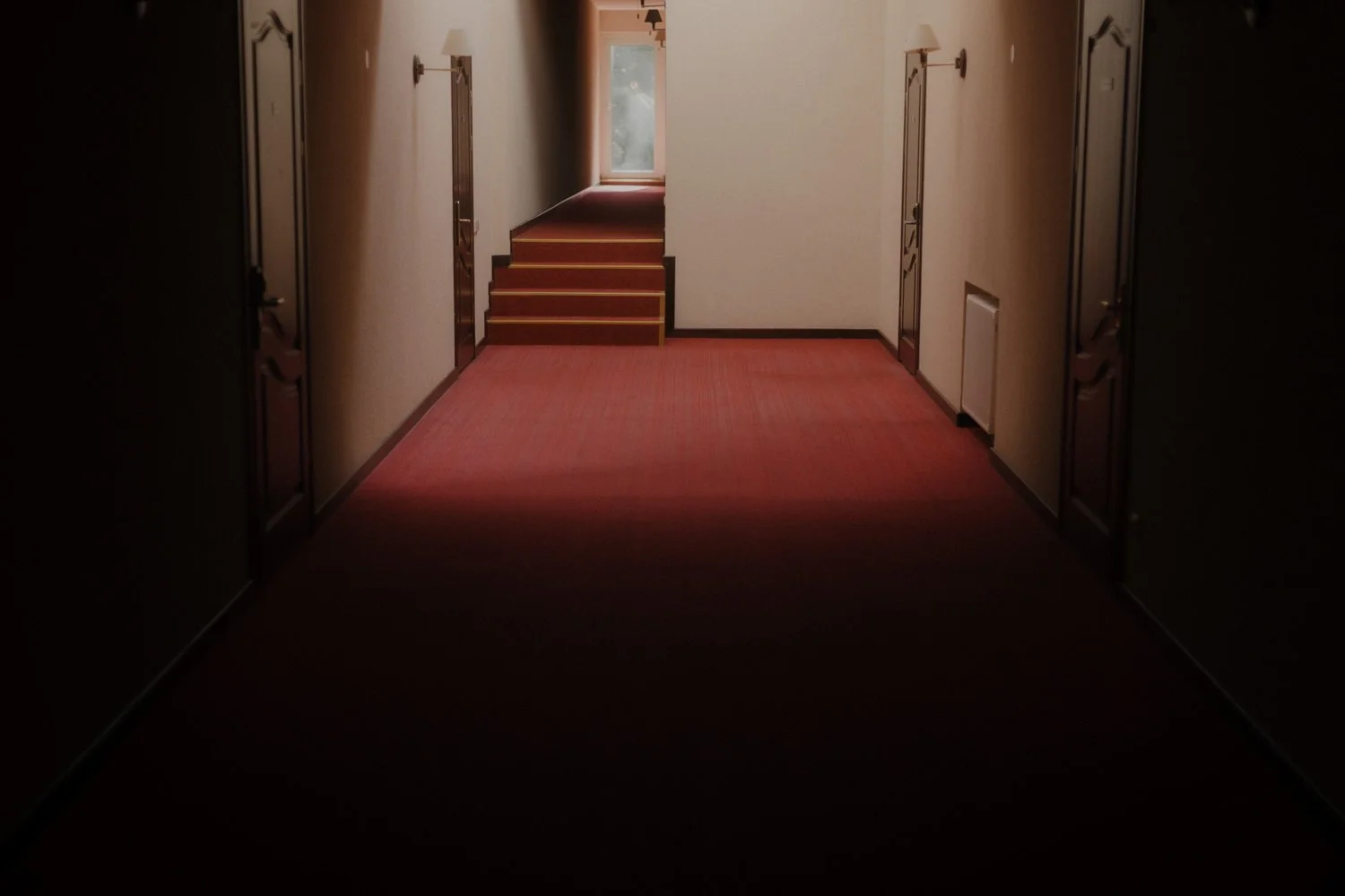 A dimly lit hotel corridor with closed wooden doors on both sides, red carpet flooring, and a staircase at the far end leading to a brightly lit area with a window.