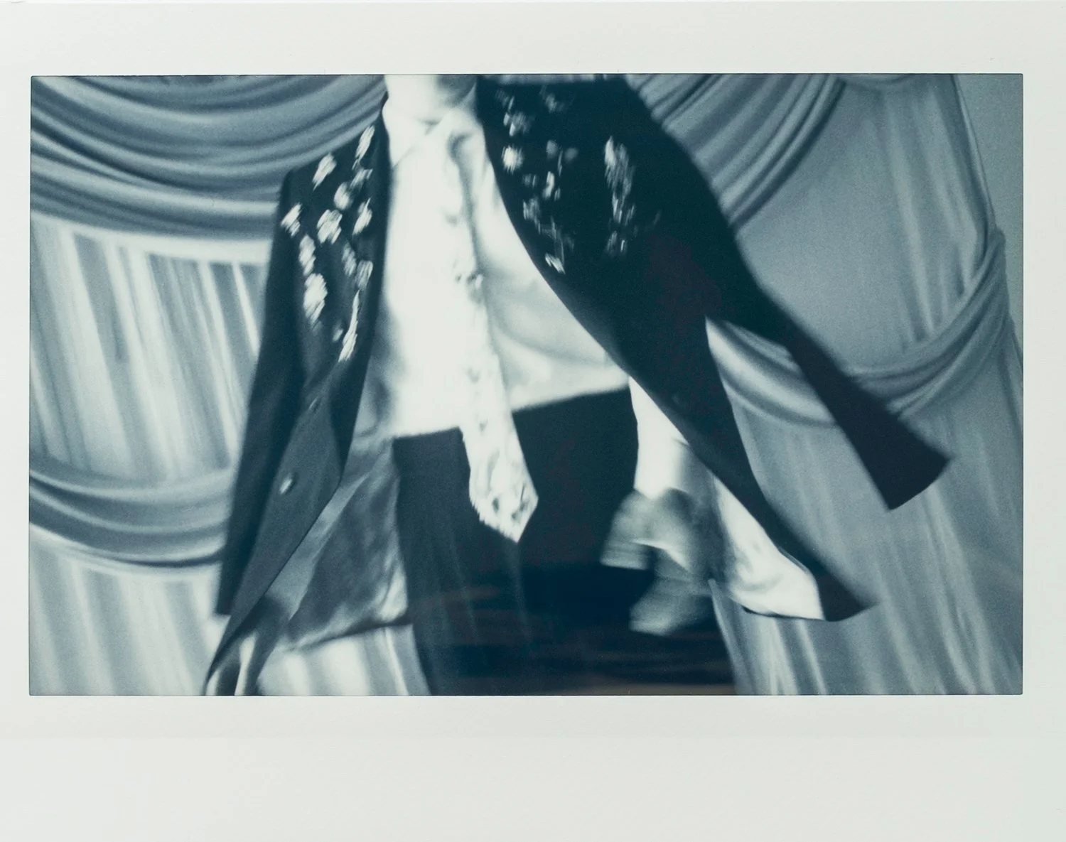 Black-and-white photo of a person wearing a scarf, open shirt, and pants, walking or running on a stage with draped curtains in the background.