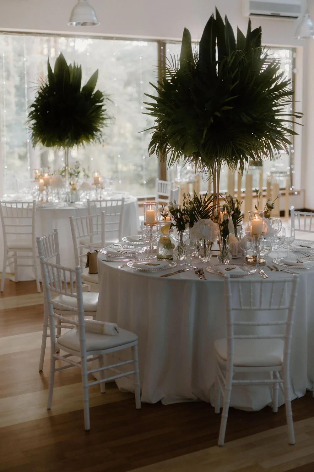 Elegant indoor wedding reception table with white tablecloth, tall floral centerpieces with green foliage, surrounded by white chairs, candlelit atmosphere, large windows in the background.