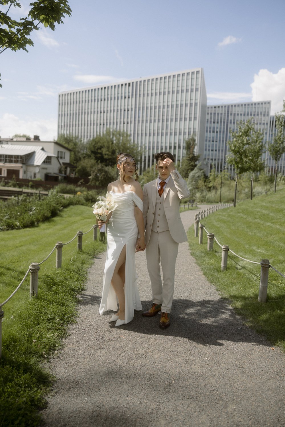 A bride and groom walking hand in hand on a gravel path in a park, with modern office buildings in the background under a partly cloudy sky.