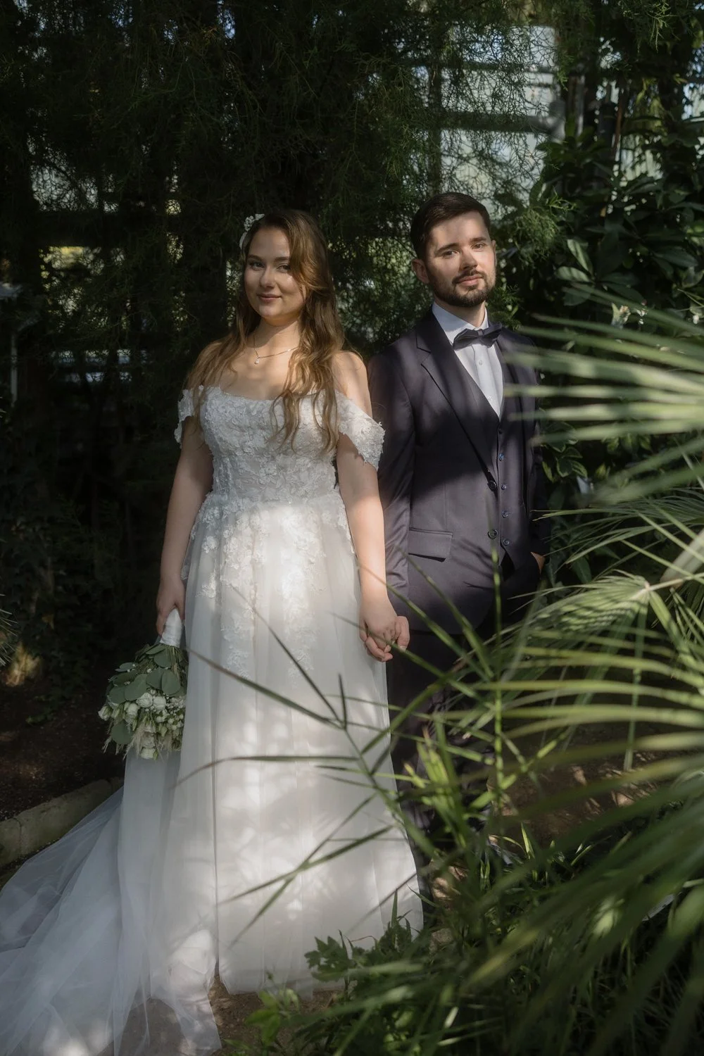 Bride and groom standing outdoors, holding hands, with lush greenery around them. The bride wears a white wedding gown and holds a bouquet, while the groom wears a dark suit with a bow tie.