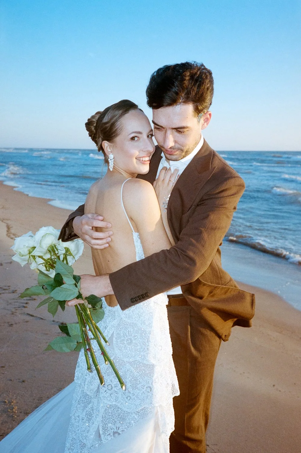 A newlywed couple hugging on the beach, the bride holding a bouquet of white roses, the groom in a brown suit, ocean waves in the background.