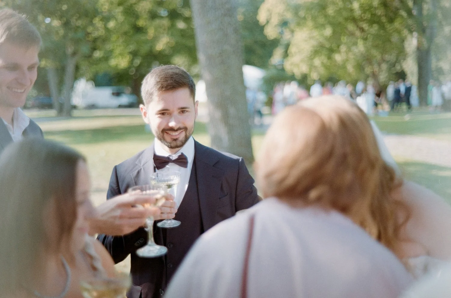 People in formal attire enjoying drinks outdoors at a social gathering in a park.