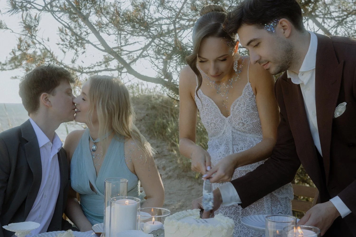 A group of people at a wedding celebration outdoors, including a couple kissing, and a bride and groom cutting a cake.