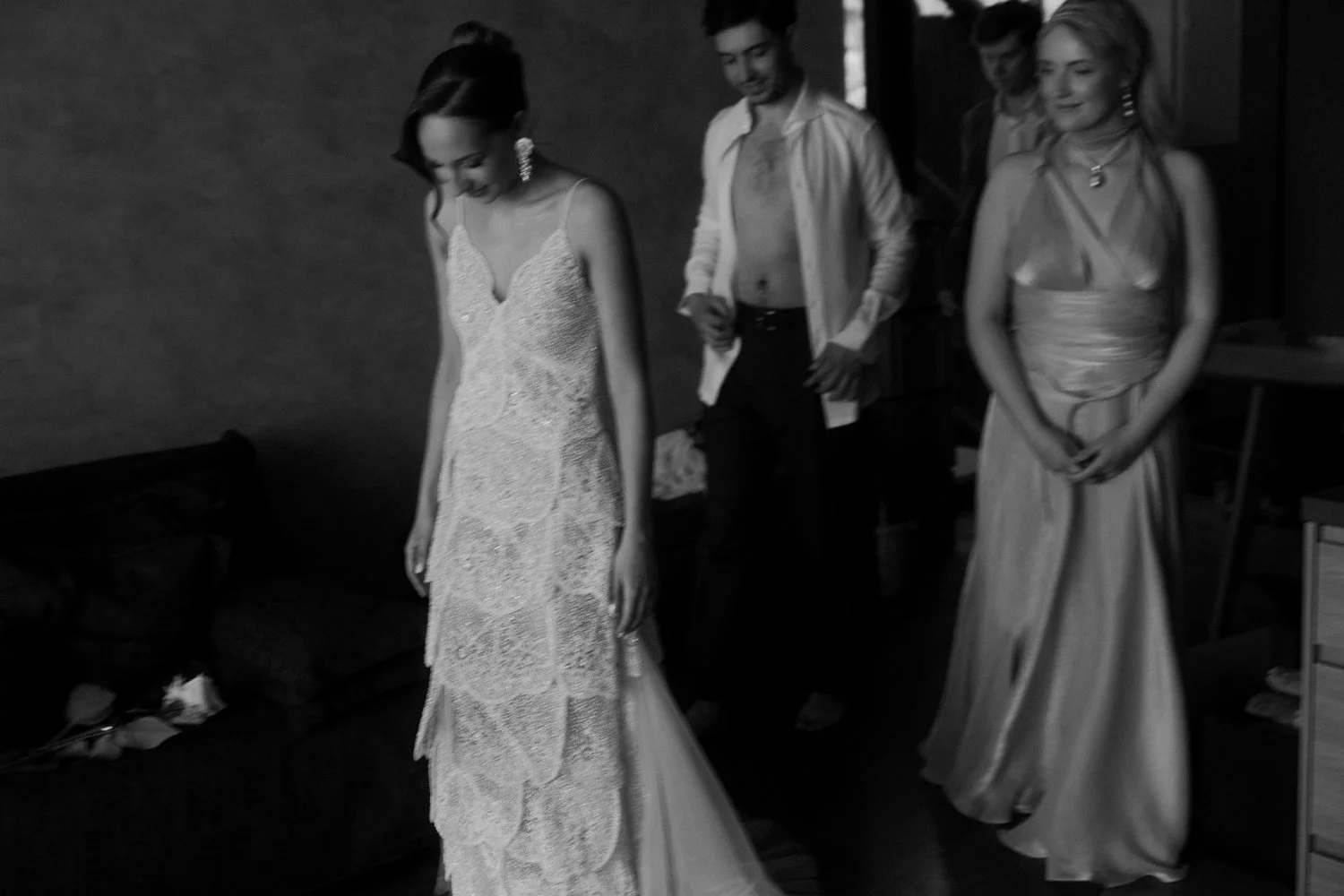 Black and white photo of three women in elegant dresses and a man with an open shirt, preparing for a formal event in a room.