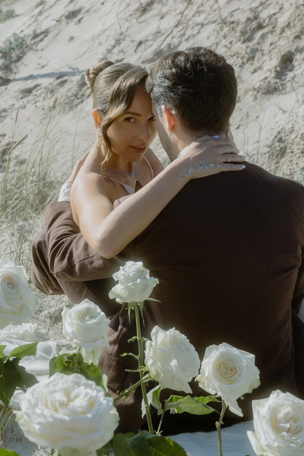 A woman and a man embrace outdoors among white roses, with a sandy hillside in the background. The woman looks into the camera, wearing a white dress and jewelry, while the man has his back to the camera.