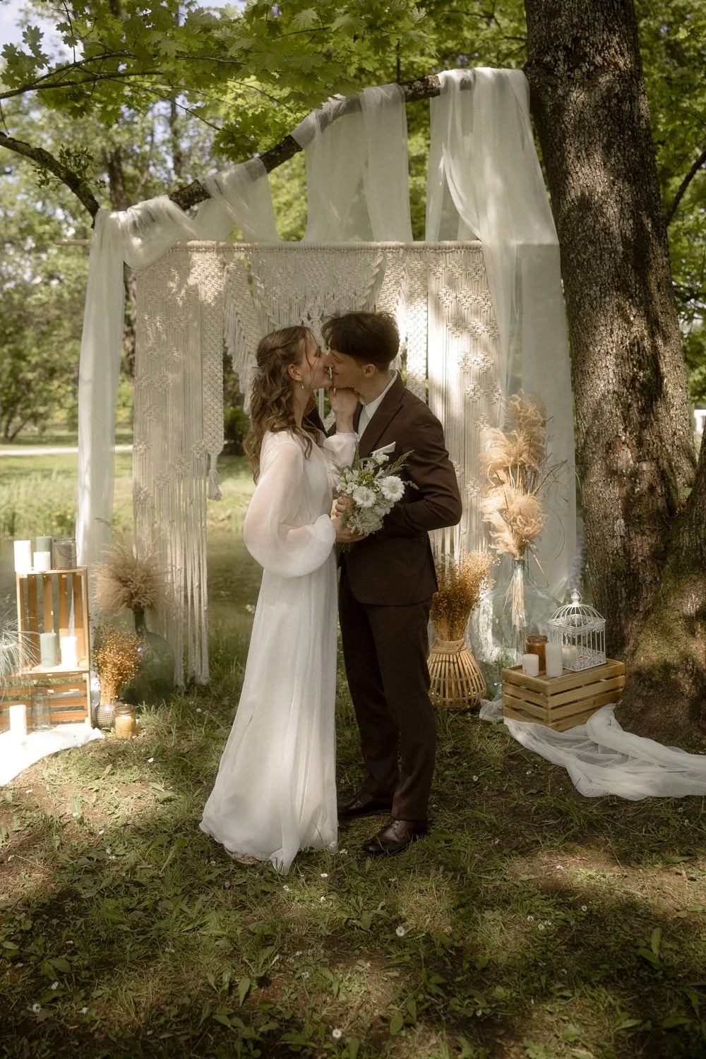 A couple in wedding attire kissing under a macrame wedding backdrop outdoors, surrounded by greenery and decorative plants, with candles and lanterns around.