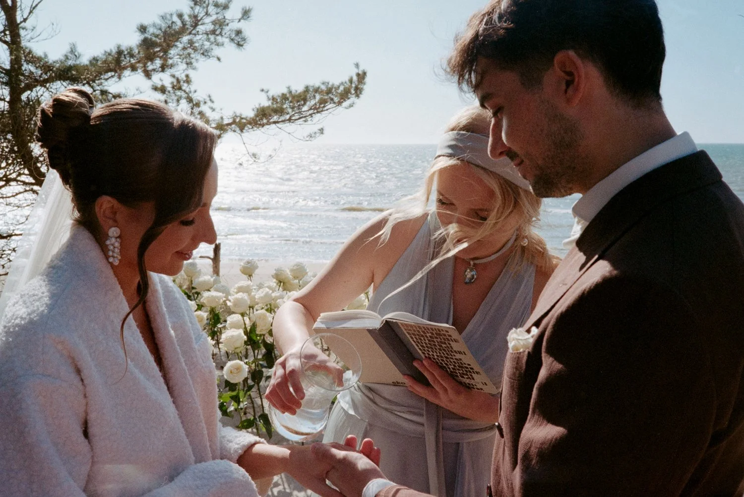 A couple getting married on a beach with an officiant, holding hands and exchanging vows, under a tree with ocean in the background.
