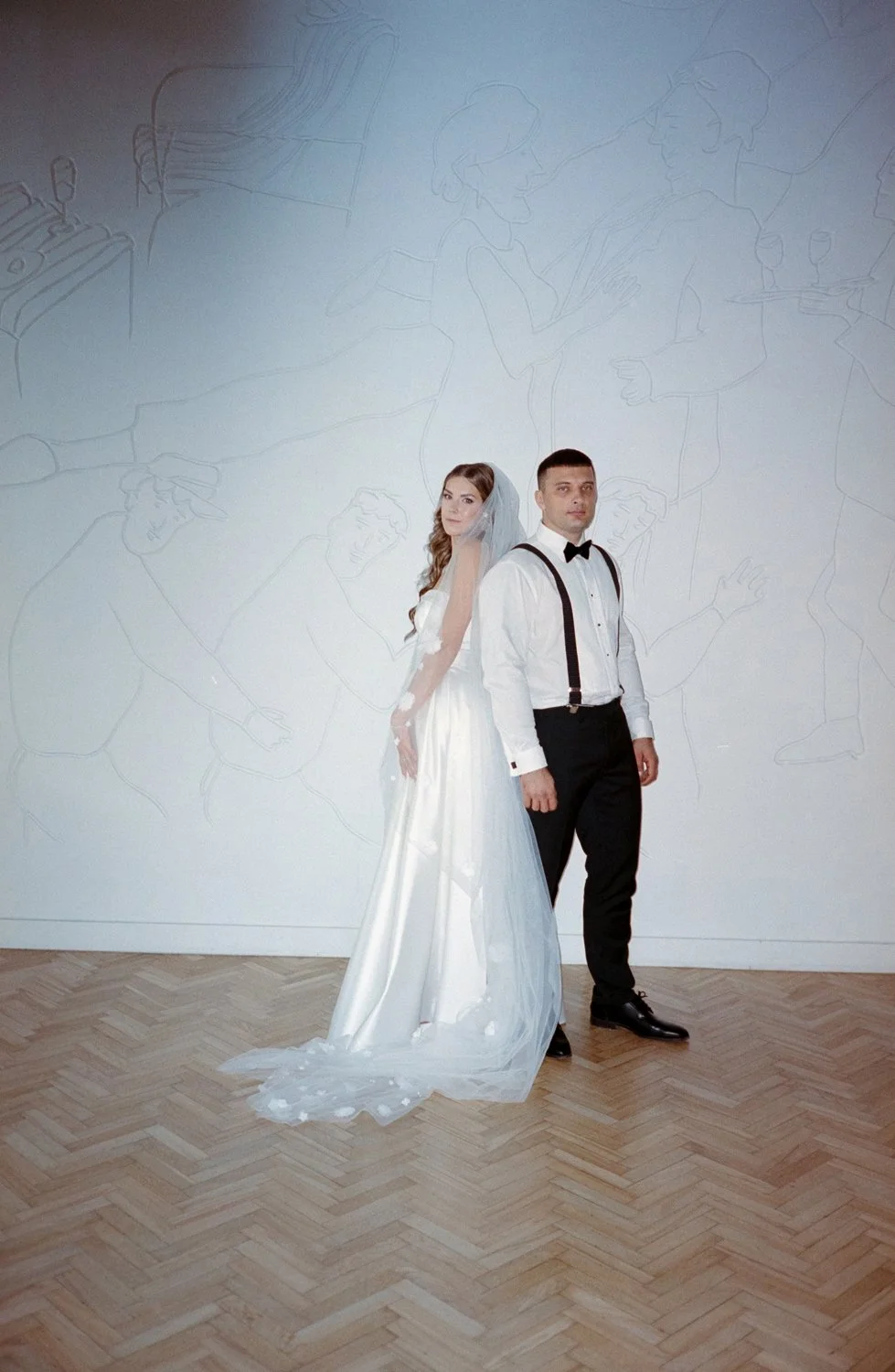 A bride and groom standing back-to-back in front of a white wall with line art drawings of people, in a modern wedding setting.