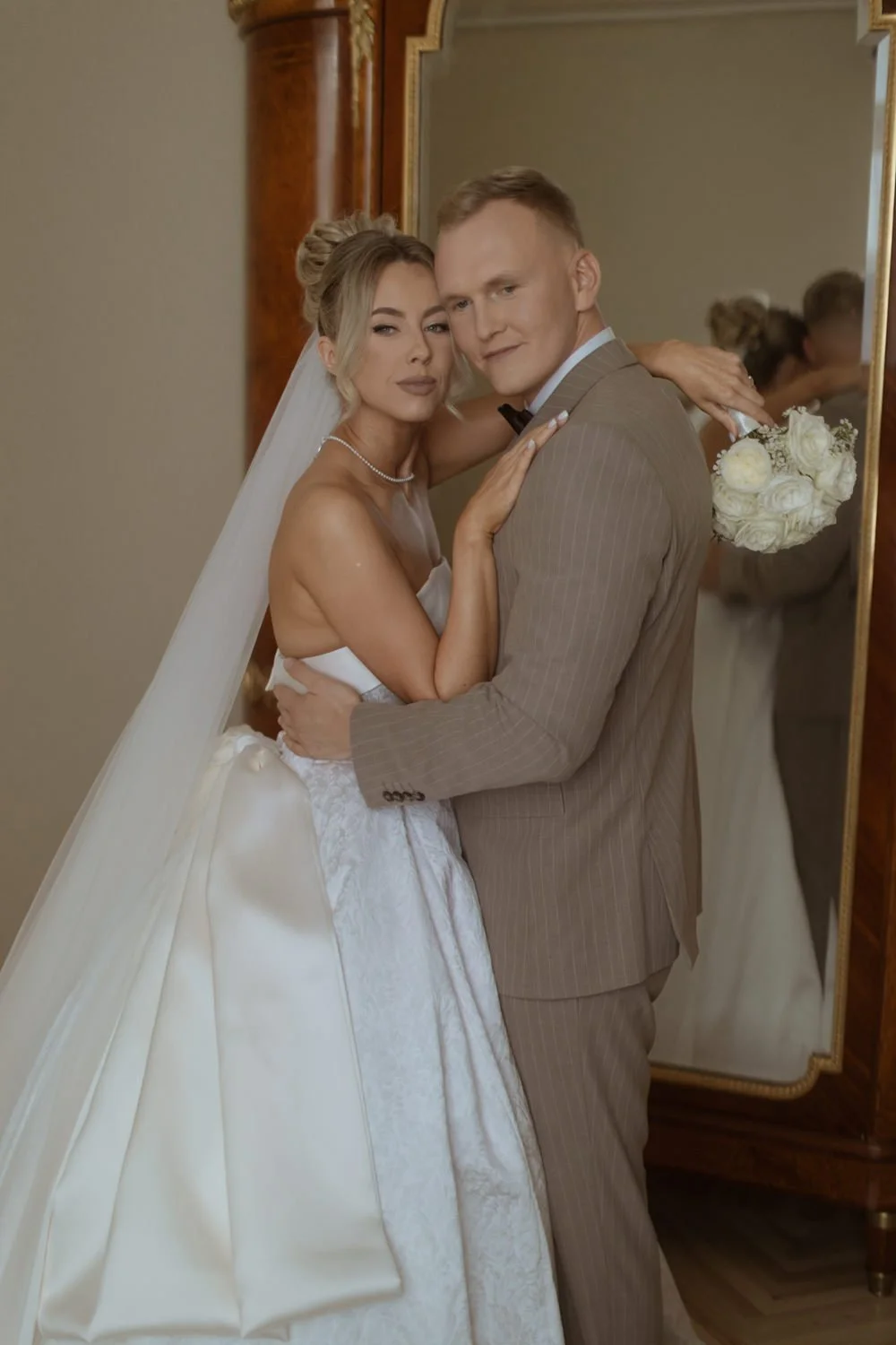 A bride and groom posing together indoors, with the bride wearing a white wedding gown and veil, and the groom in a brown striped suit. The bride is holding a bouquet of white flowers and has her arm around the groom's shoulder, both smiling softly.