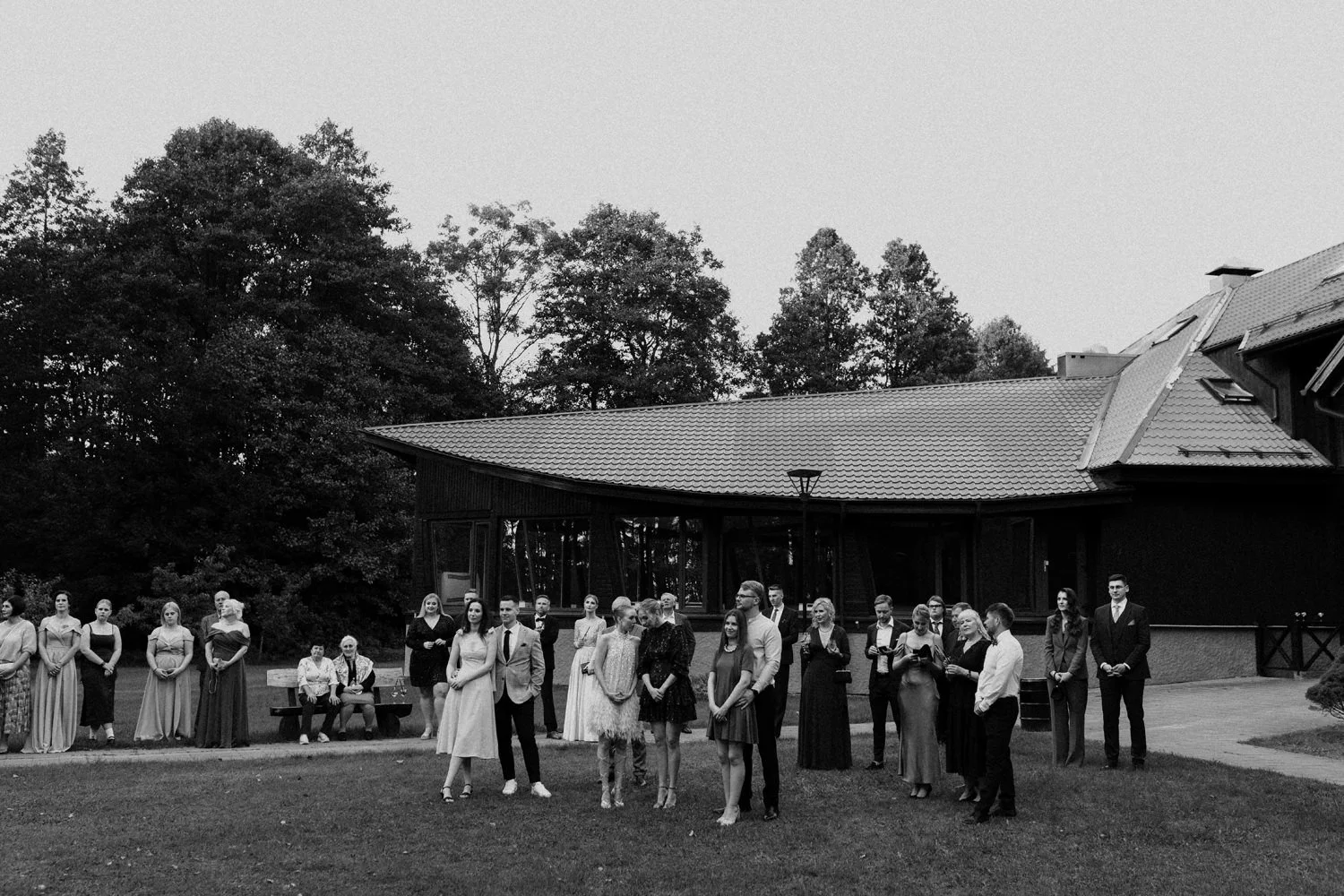 Black and white photo of a group of people dressed formally, standing on a lawn in front of a modern house with large windows, surrounded by trees.
