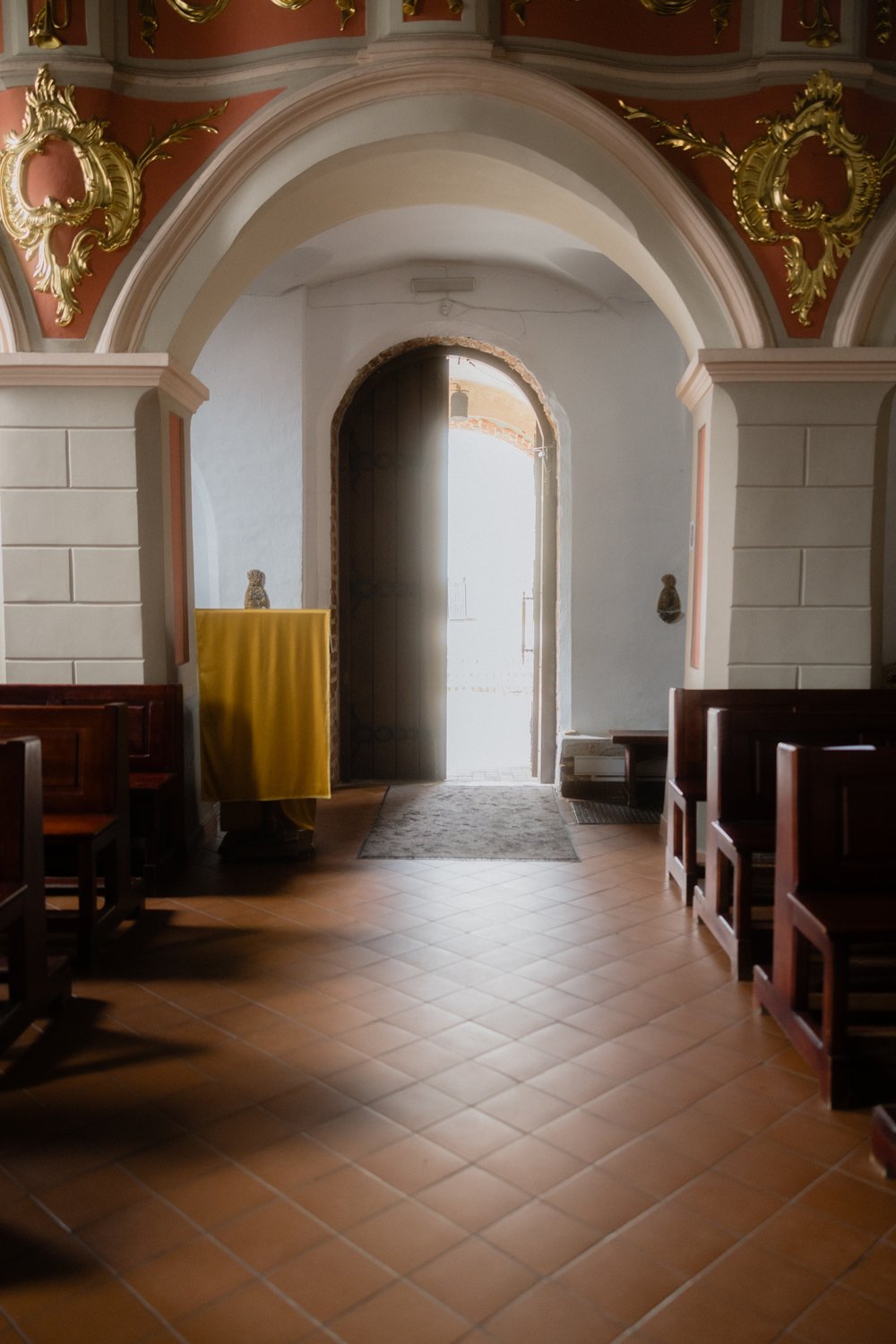 Interior of a church or chapel with wooden pews, ornate gold embellishments on the archway, and an open door letting in natural light.