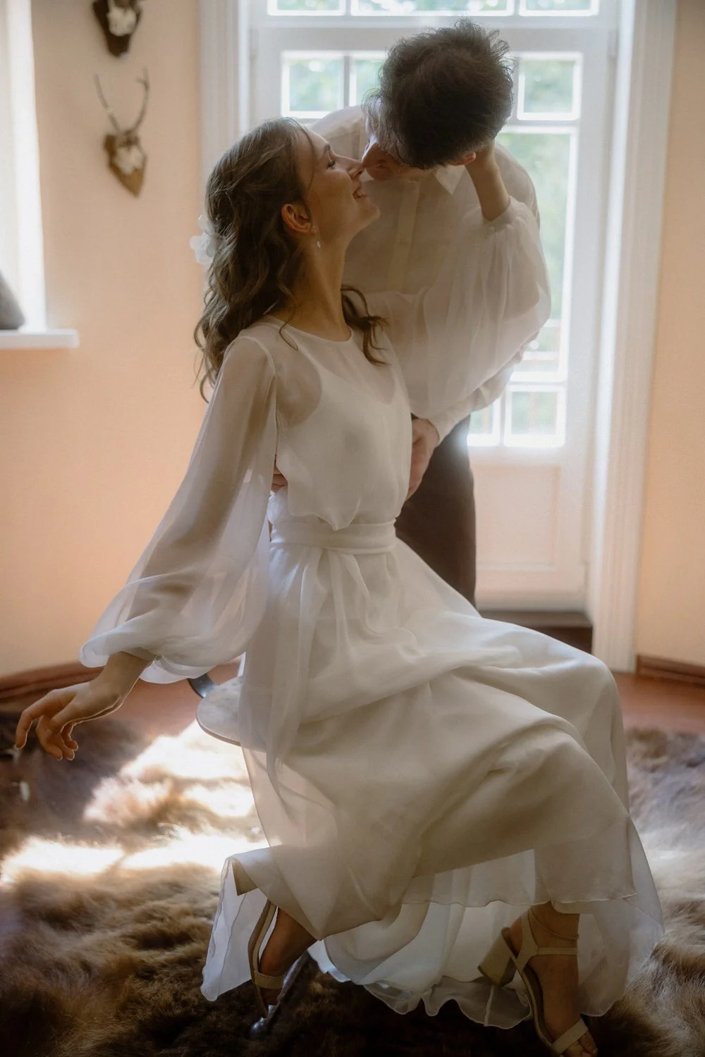 A woman in a white dress sitting on a furry rug, leaning back while a man leaning toward her, both sharing a kiss indoors near a window.