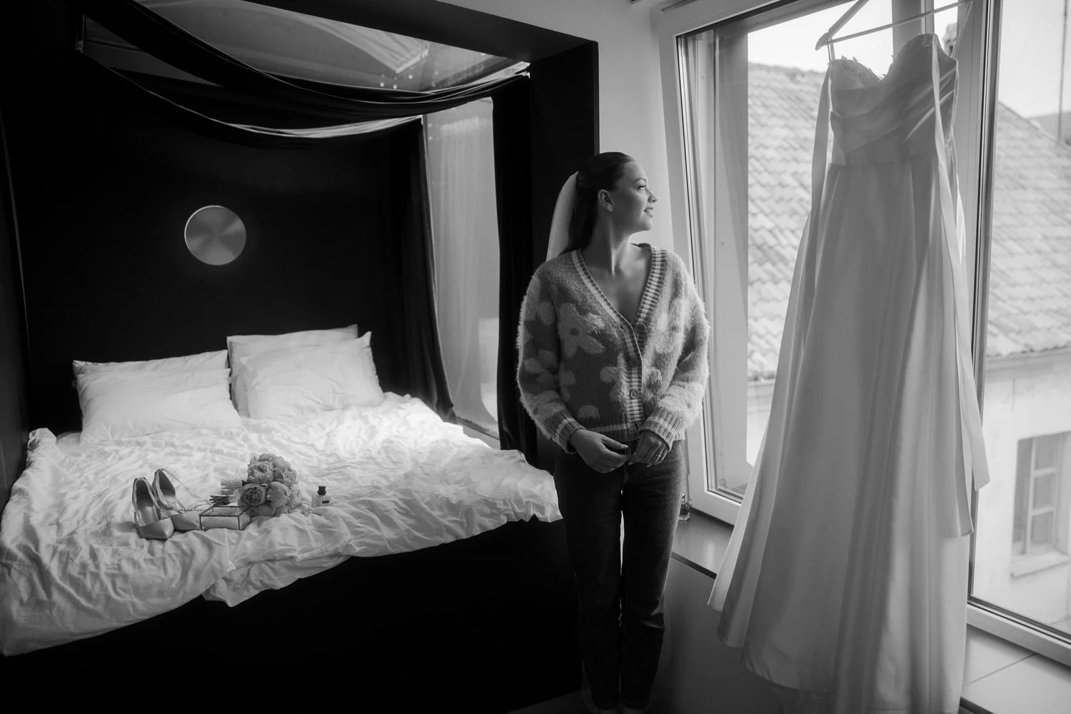 A woman stands by a window in a bedroom, looking at a wedding dress hanging outside the window. The room has a bed with pillows, and on the bed are bridal shoes, a bouquet, and small boxes.