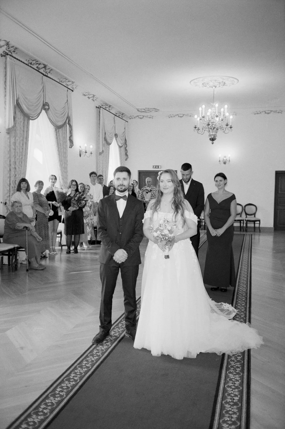 Black and white photo of a wedding ceremony with a bride in a wedding gown holding a bouquet and a groom in a suit standing at the front of a decorated hall with guests in the background.