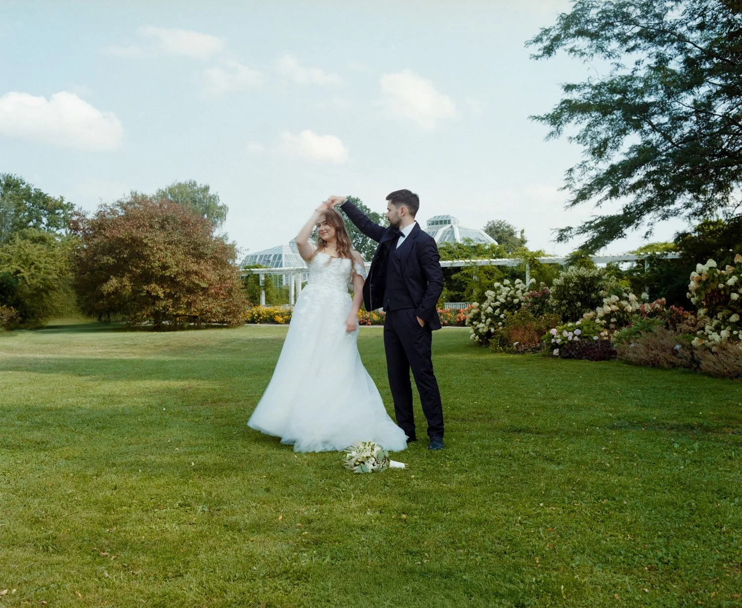 A bride and groom dance outdoors on a grassy lawn with trees, flowers, and a greenhouse in the background.