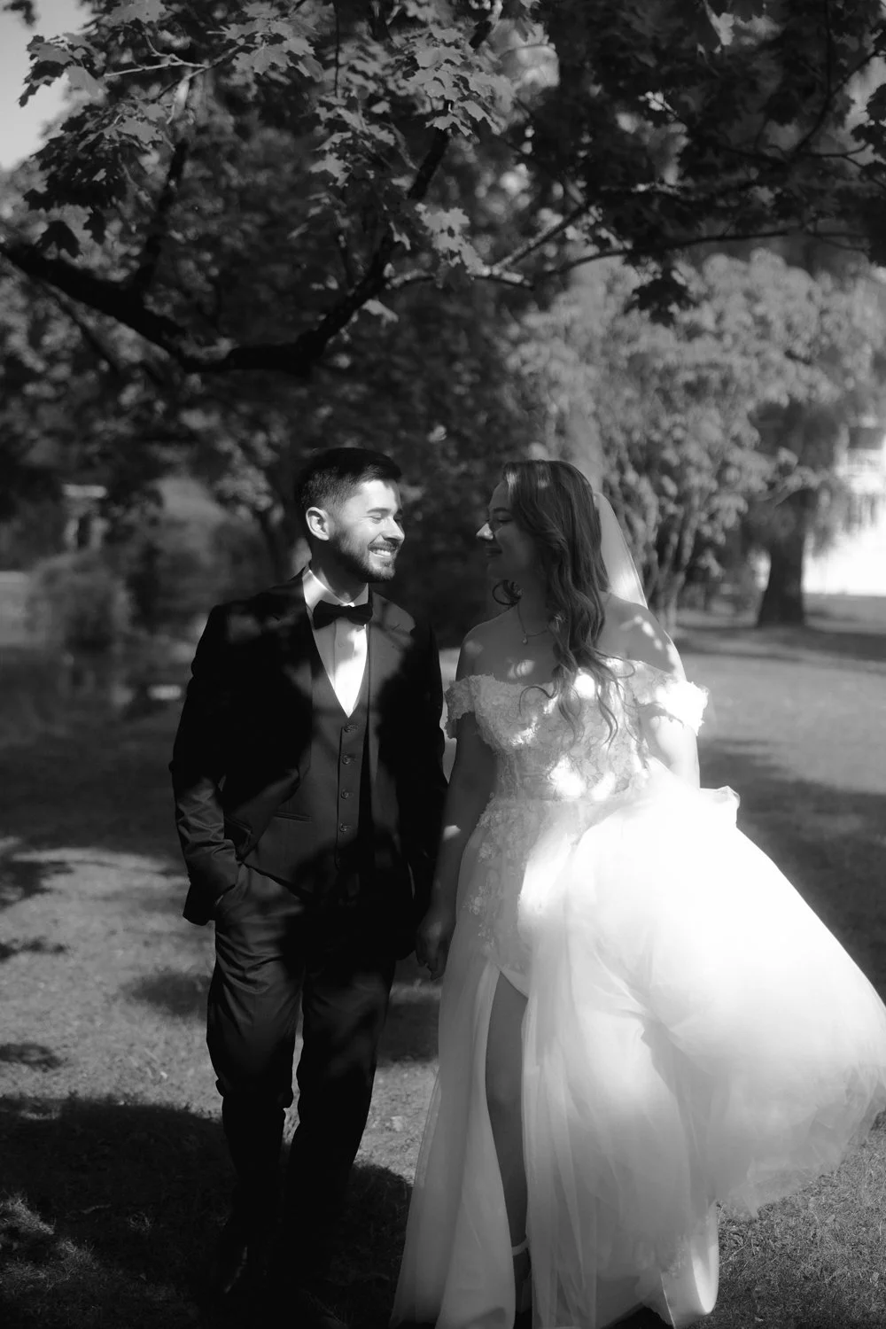 A bride and groom are walking outdoors, holding hands, and smiling at each other on their wedding day, with trees and sunlight in the background.