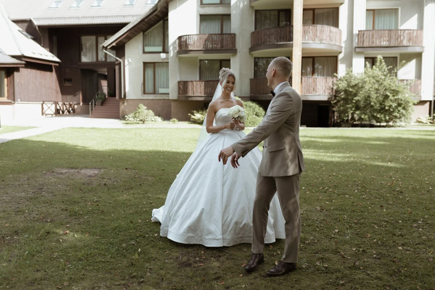 A bride in a white wedding dress holding a bouquet, smiling at a groom in a tan suit, outside on a grassy area in front of a multi-story residential building with balconies.