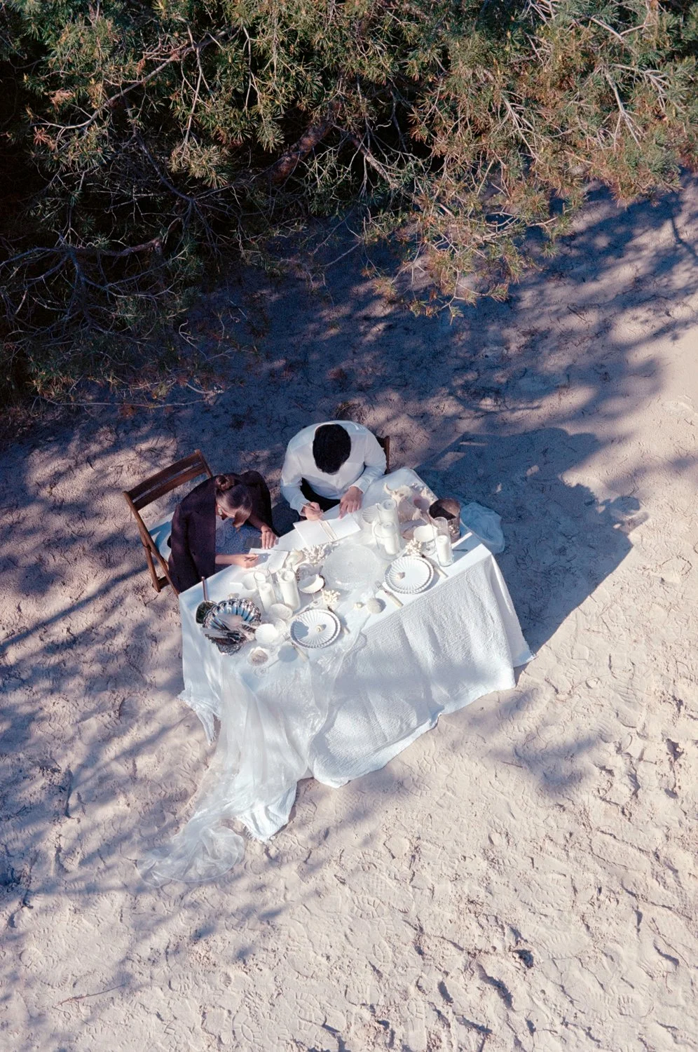 People having a meal outdoors at a table covered with a white cloth, set with dishes and utensils, next to a tree on sandy ground, viewed from above.
