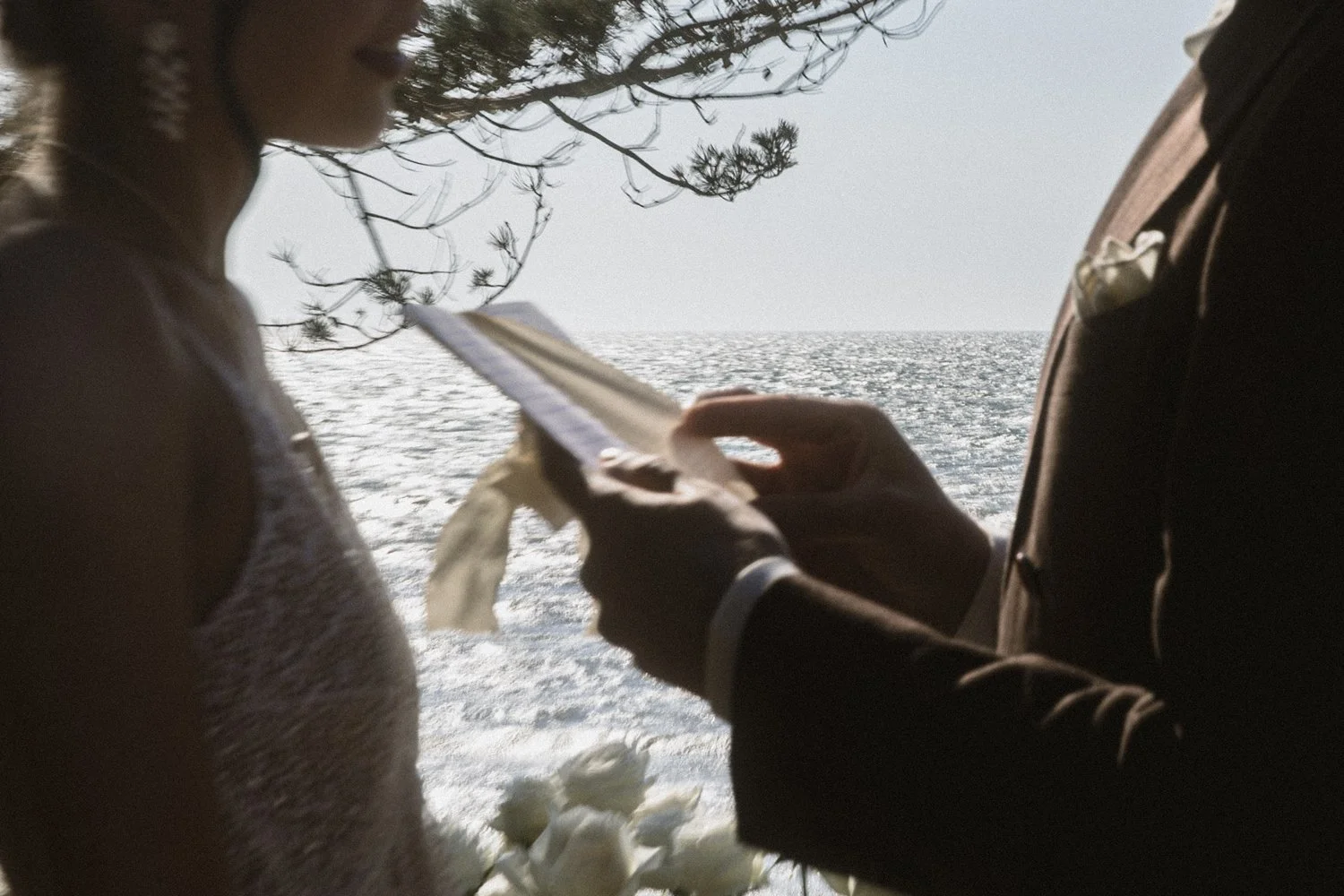 A couple exchanging vows at a beach wedding by the ocean, with the bride and groom holding a ring and flower petals against a backdrop of the sea and clouds.