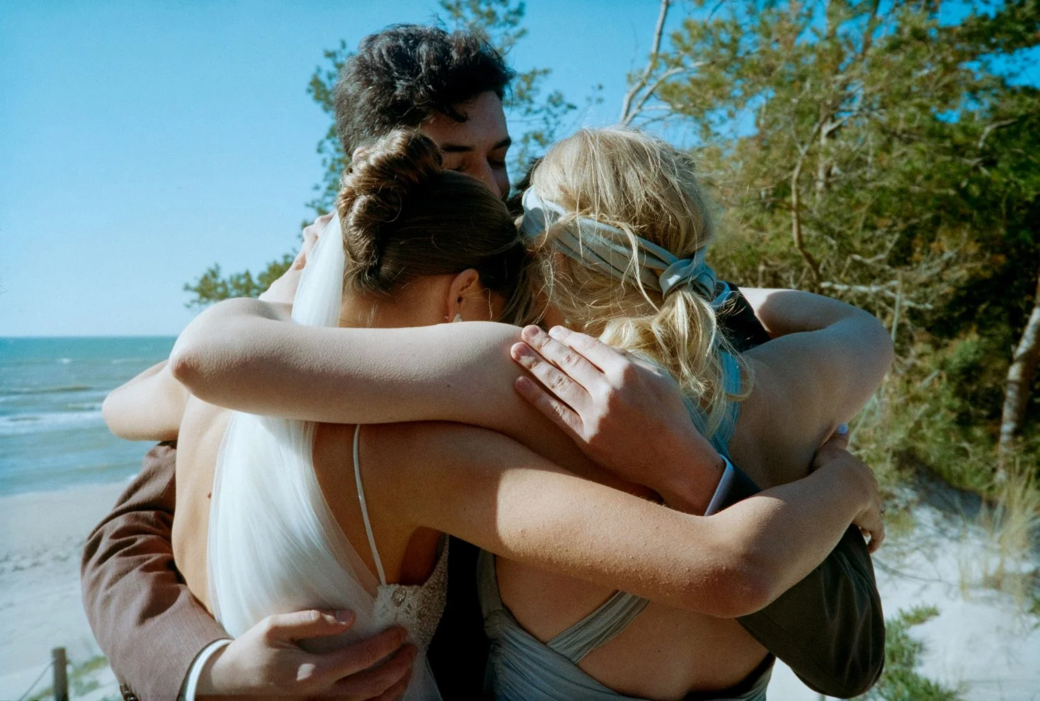 Four people hugging tightly on a beach with trees and ocean in the background.