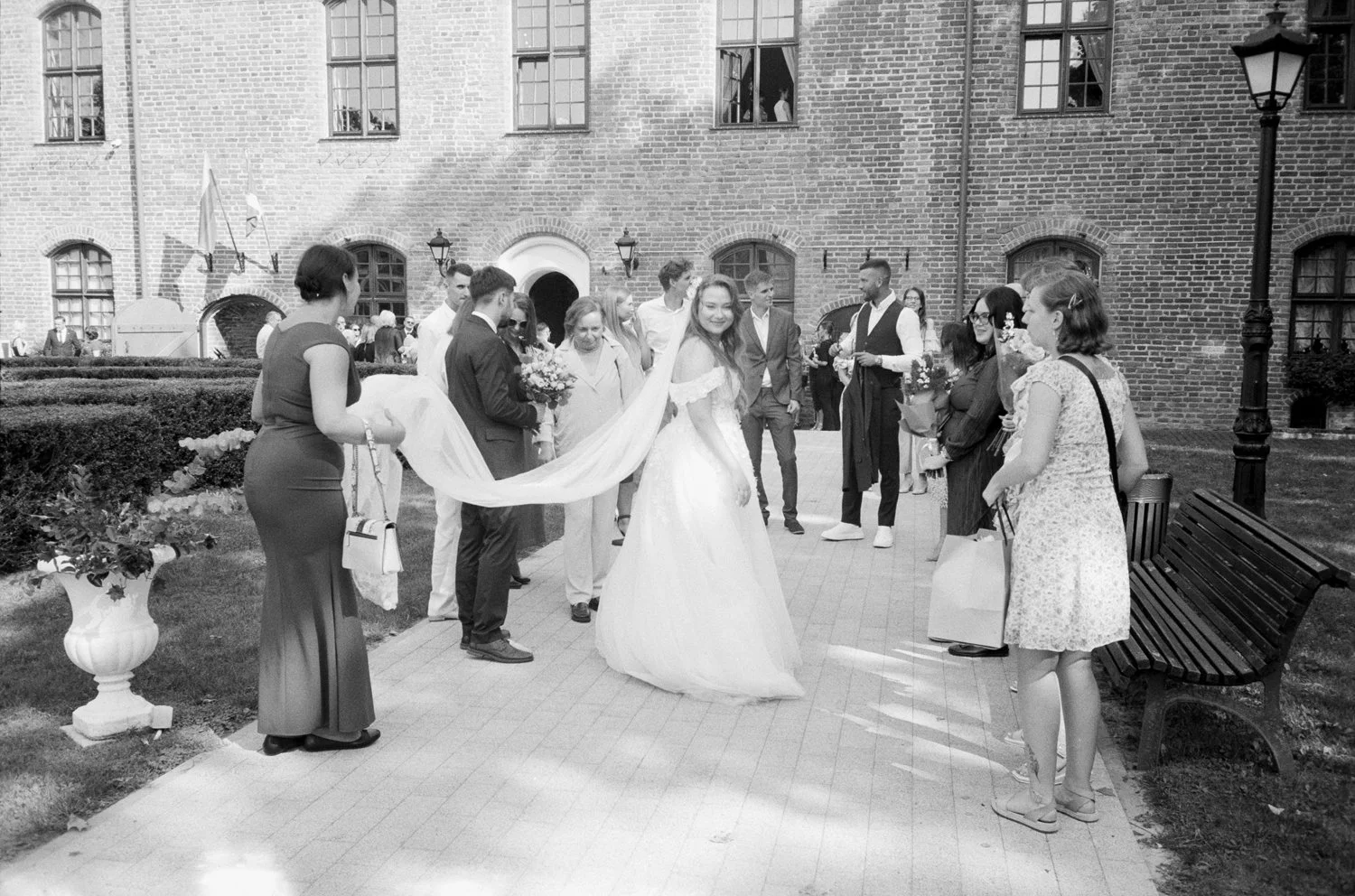 A group of people gathered outside a brick building, with a woman in a wedding dress in the center, surrounded by friends and family holding flowers and smiling