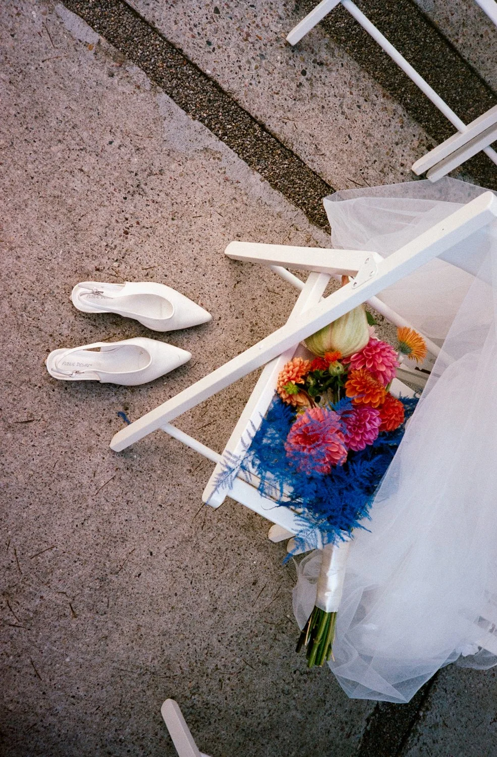 White high-heeled shoes, a bouquet of colorful flowers, and a white chair on a concrete surface.