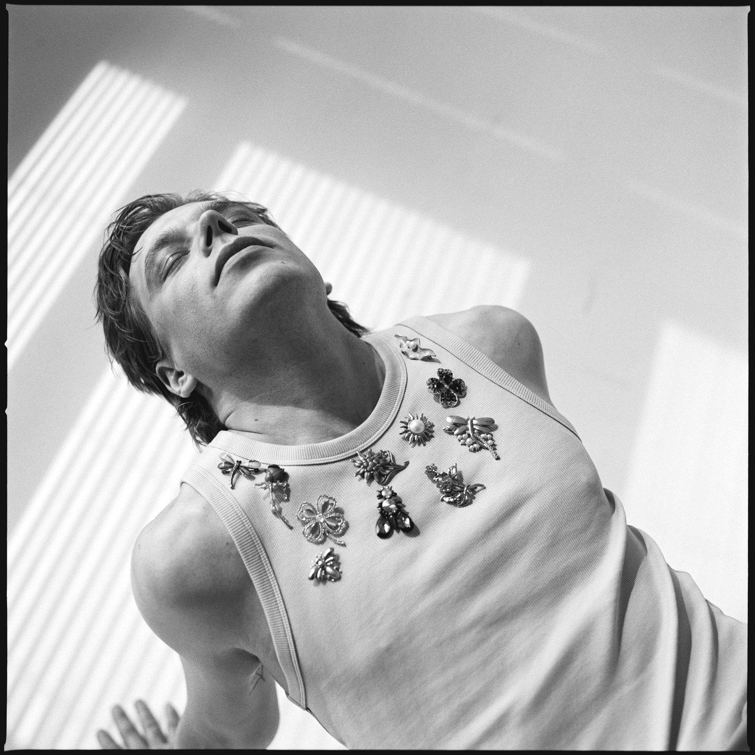 A black-and-white image of a woman with short hair, eyes closed, looking upward with a peaceful expression, wearing a sleeveless top adorned with various decorative pins and brooches.