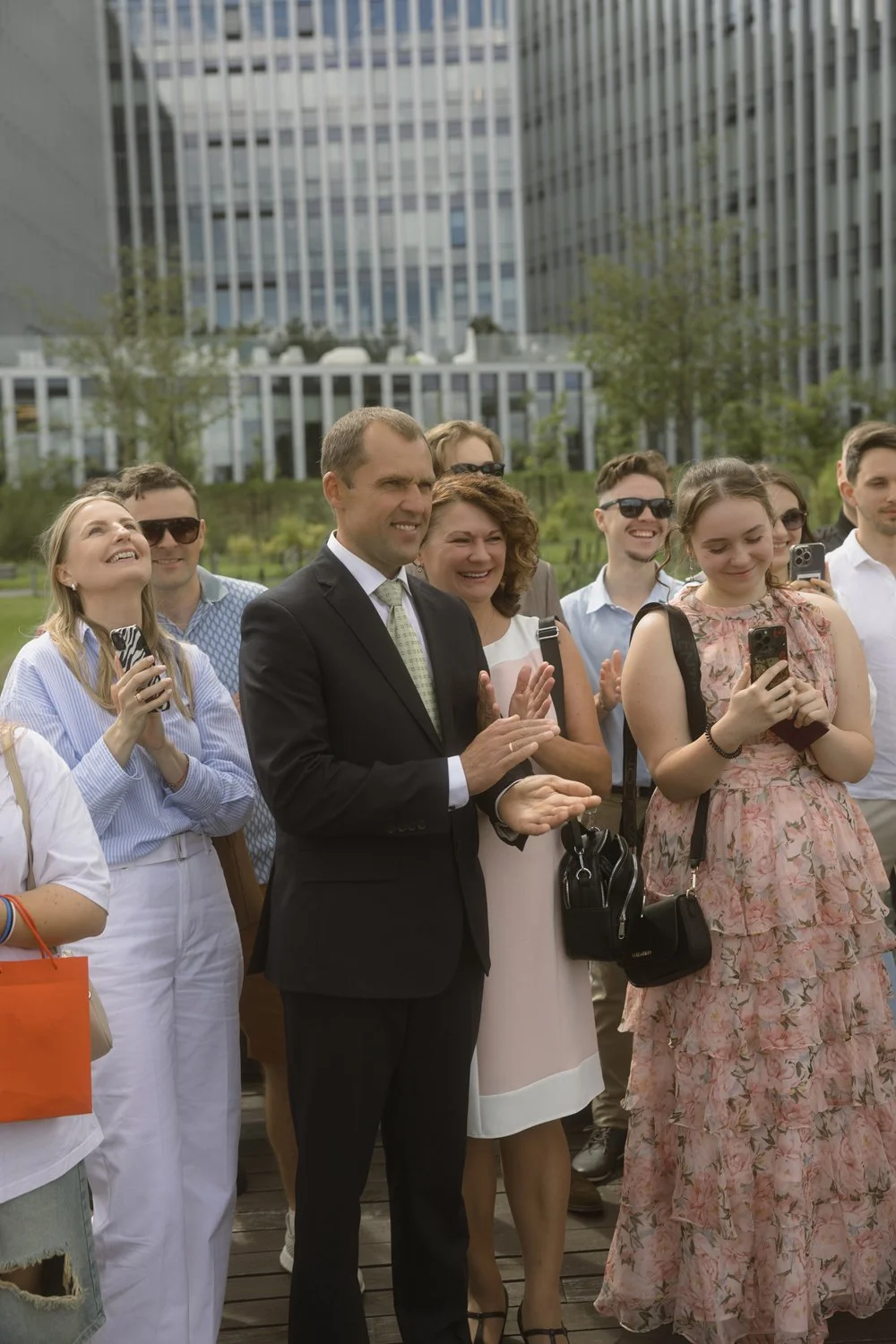 A group of people, including a man in a suit, gathered outdoors in front of modern office buildings, watching and clapping.
